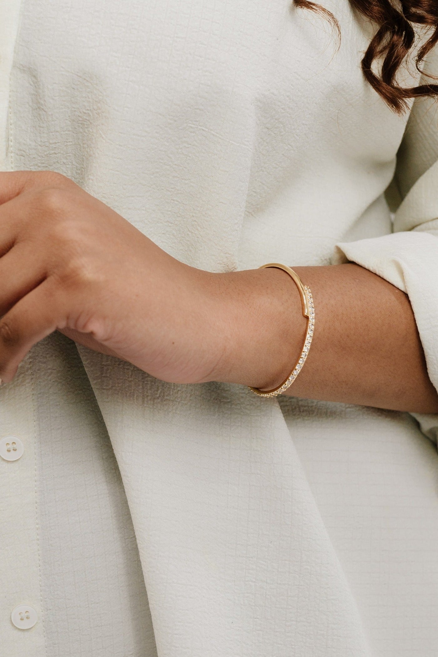 A hand is resting on a light-colored shirt, showcasing a gold bracelet adorned with sparkling stones. The shirt has a textured pattern, providing a soft, elegant backdrop.