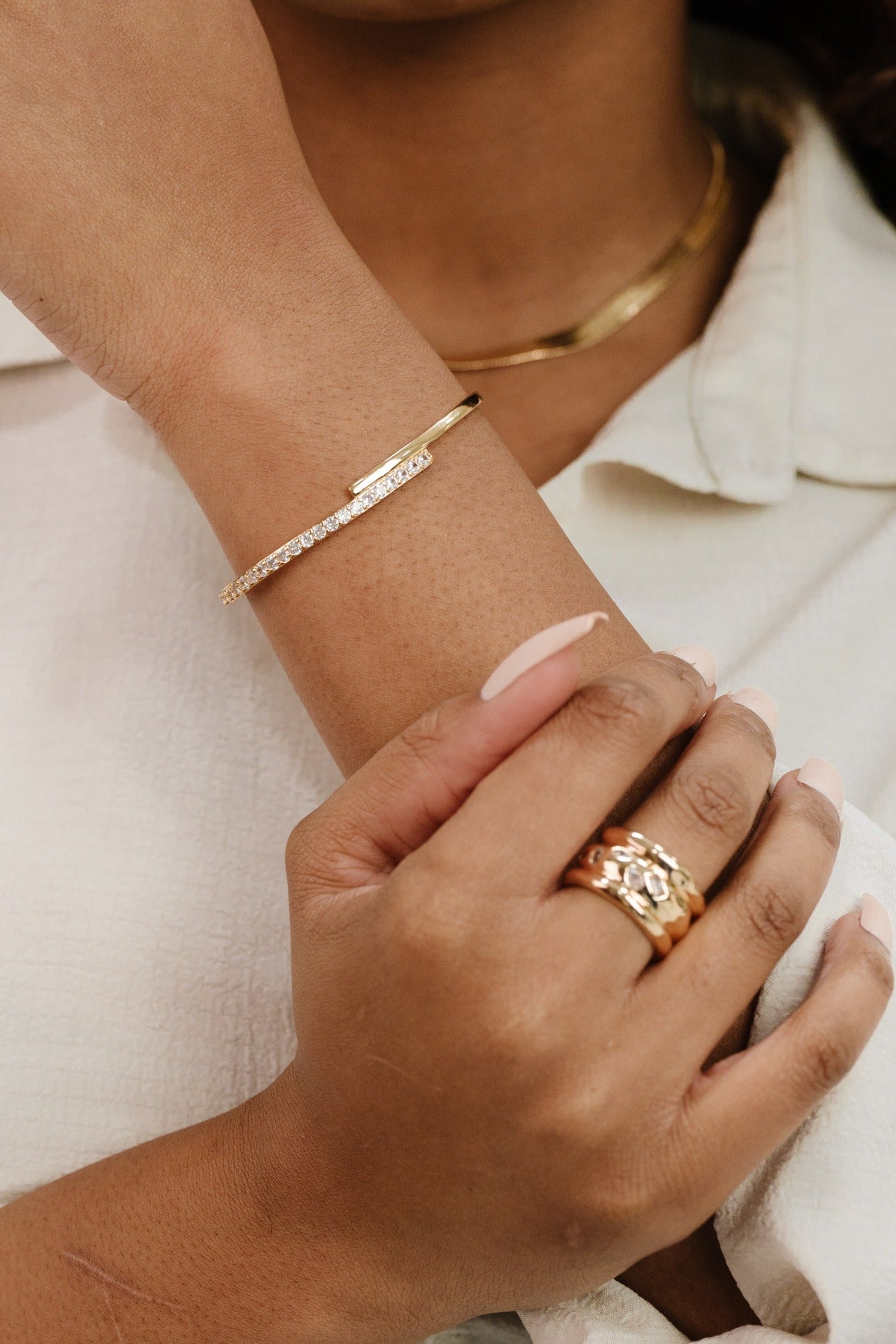 A hand adorned with multiple rings is intertwined with another wrist displaying a delicate gold bracelet. The background features a soft, neutral fabric, enhancing the elegant jewelry.