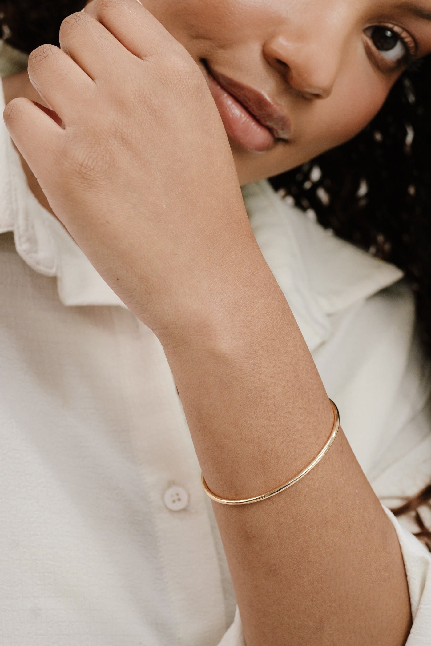 A hand with a gold bangle is gently touching the face. The person is wearing a light-colored shirt, and the setting appears soft and casual, focusing on personal style.