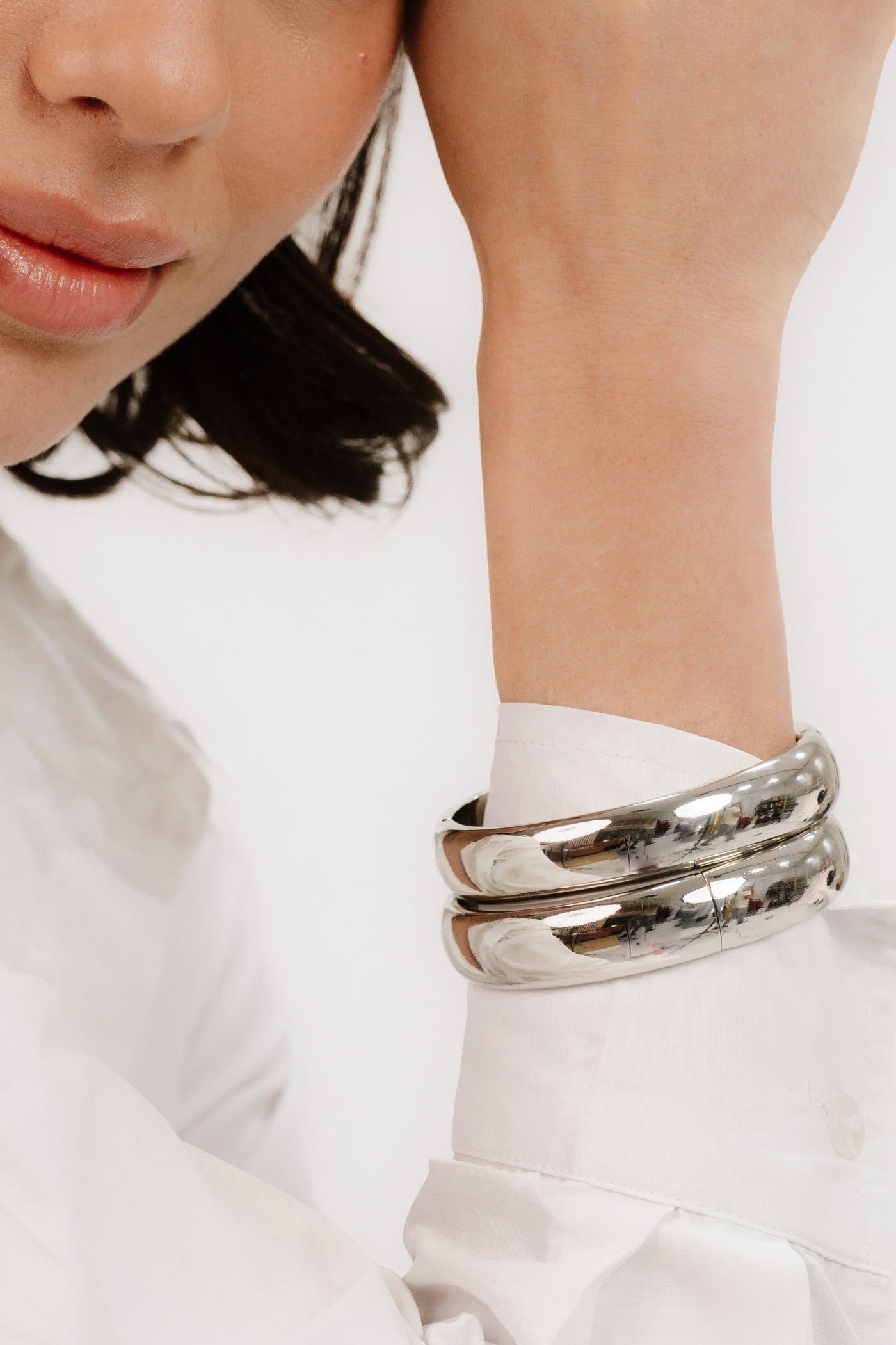 A close-up of a woman’s wrist adorned with three shiny metallic bangles, as she lightly rests her hand against her face. She wears a white, long-sleeved shirt.