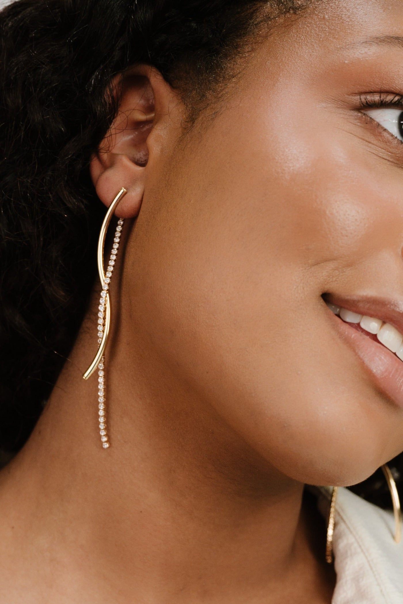 A close-up of a woman's face showcases elegant, elongated gold earrings with a dangling rhinestone chain. Her smiling expression contributes to a stylish, confident appearance against a softly blurred background.