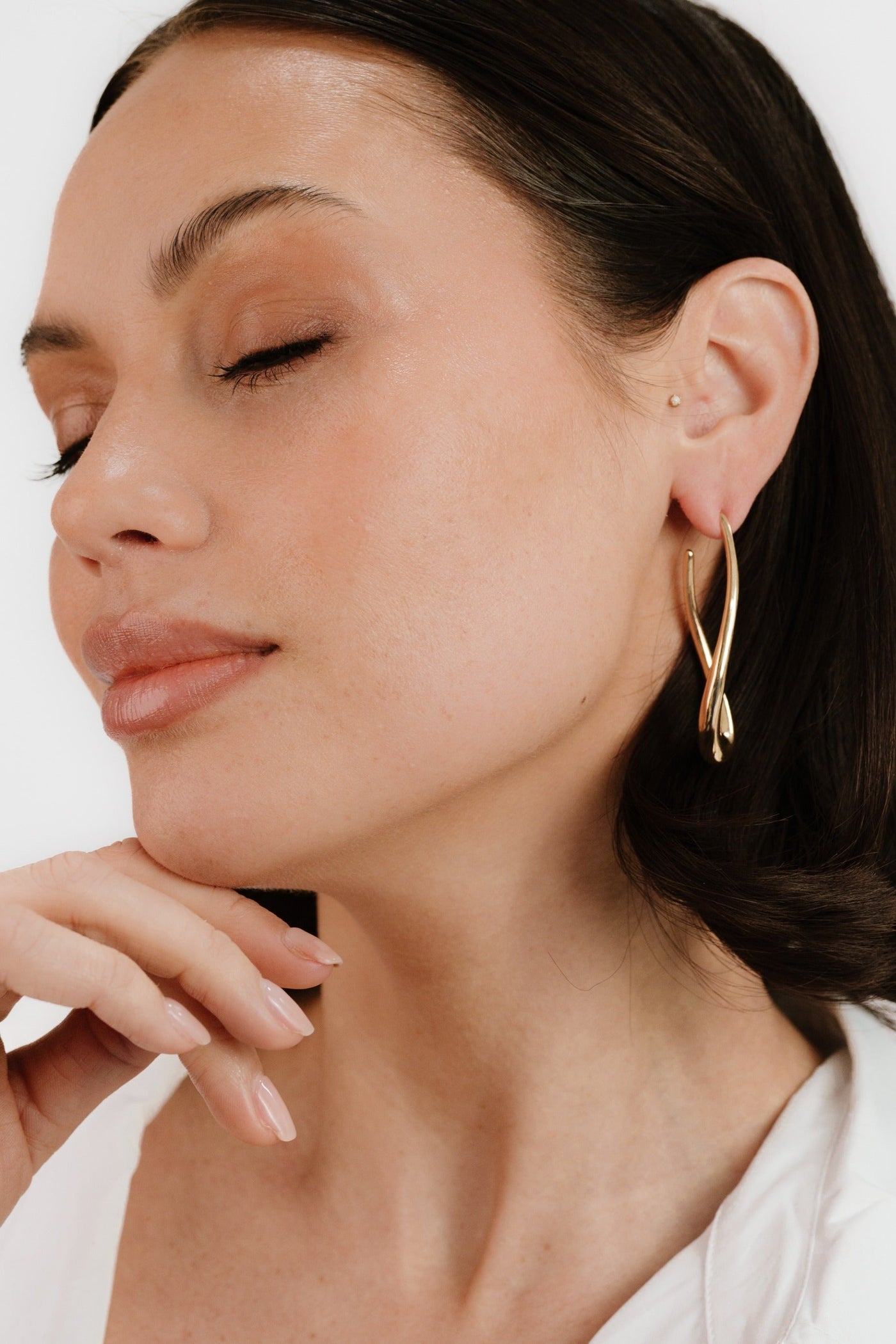 A woman with closed eyes gently rests her chin on her hand, showcasing elegant gold earrings against a plain white background. Her skin has a smooth, natural appearance.