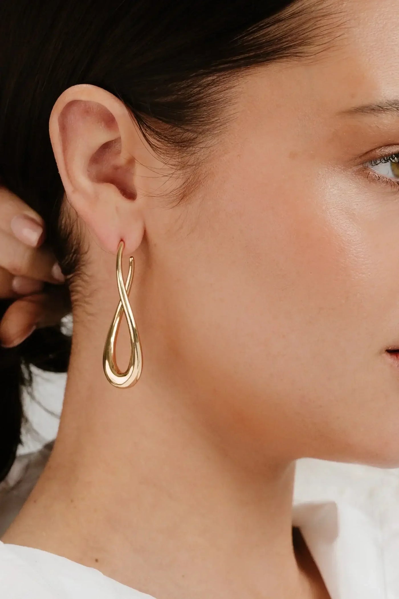 A close-up of a woman’s face highlighting a shiny, intricate gold earring dangling from her ear. She is gently touching her hair with one hand, set against a light background.