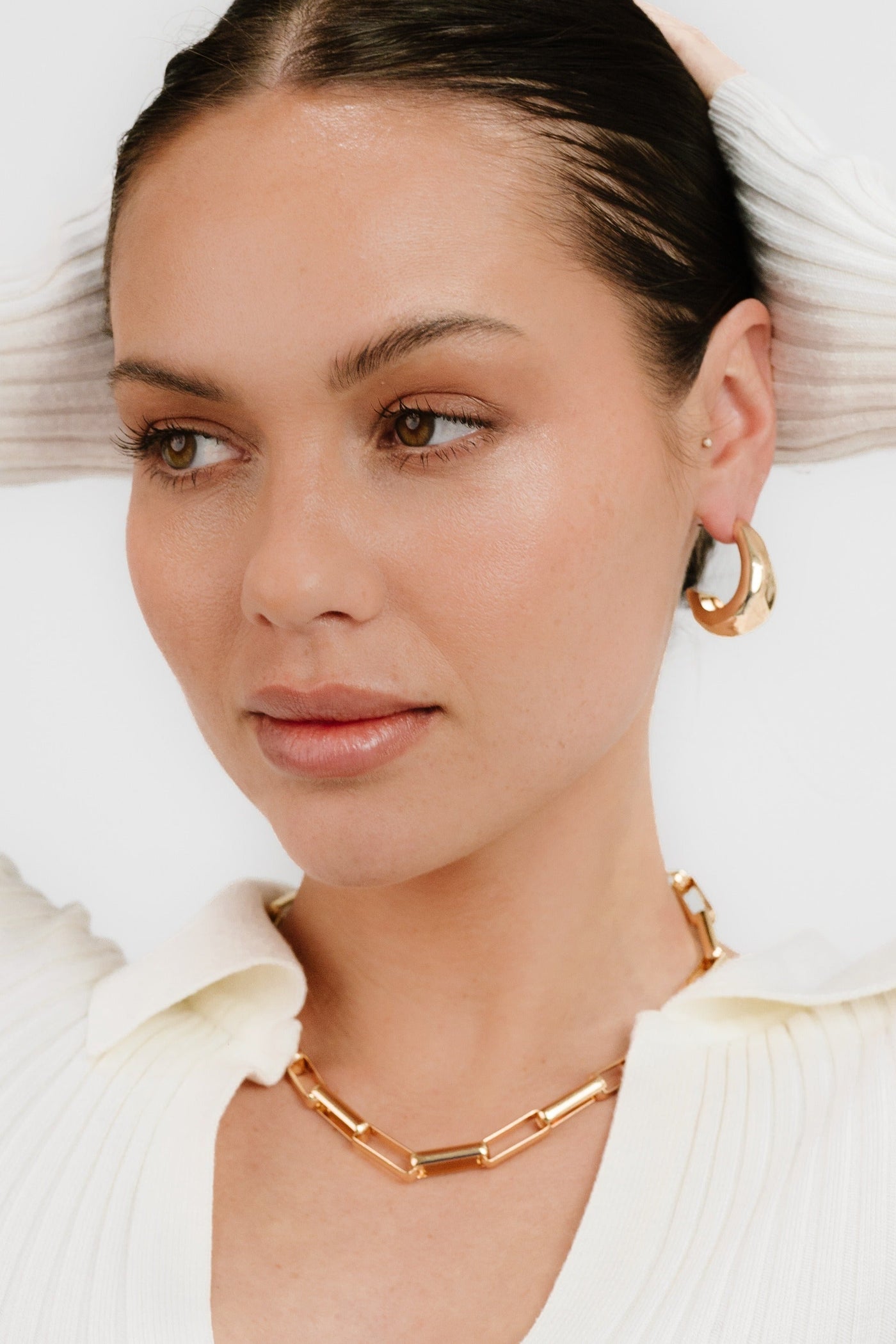 A woman with a natural makeup look is thoughtfully posed, touching her hair. She wears gold hoop earrings and a gold chain necklace, set against a light background.