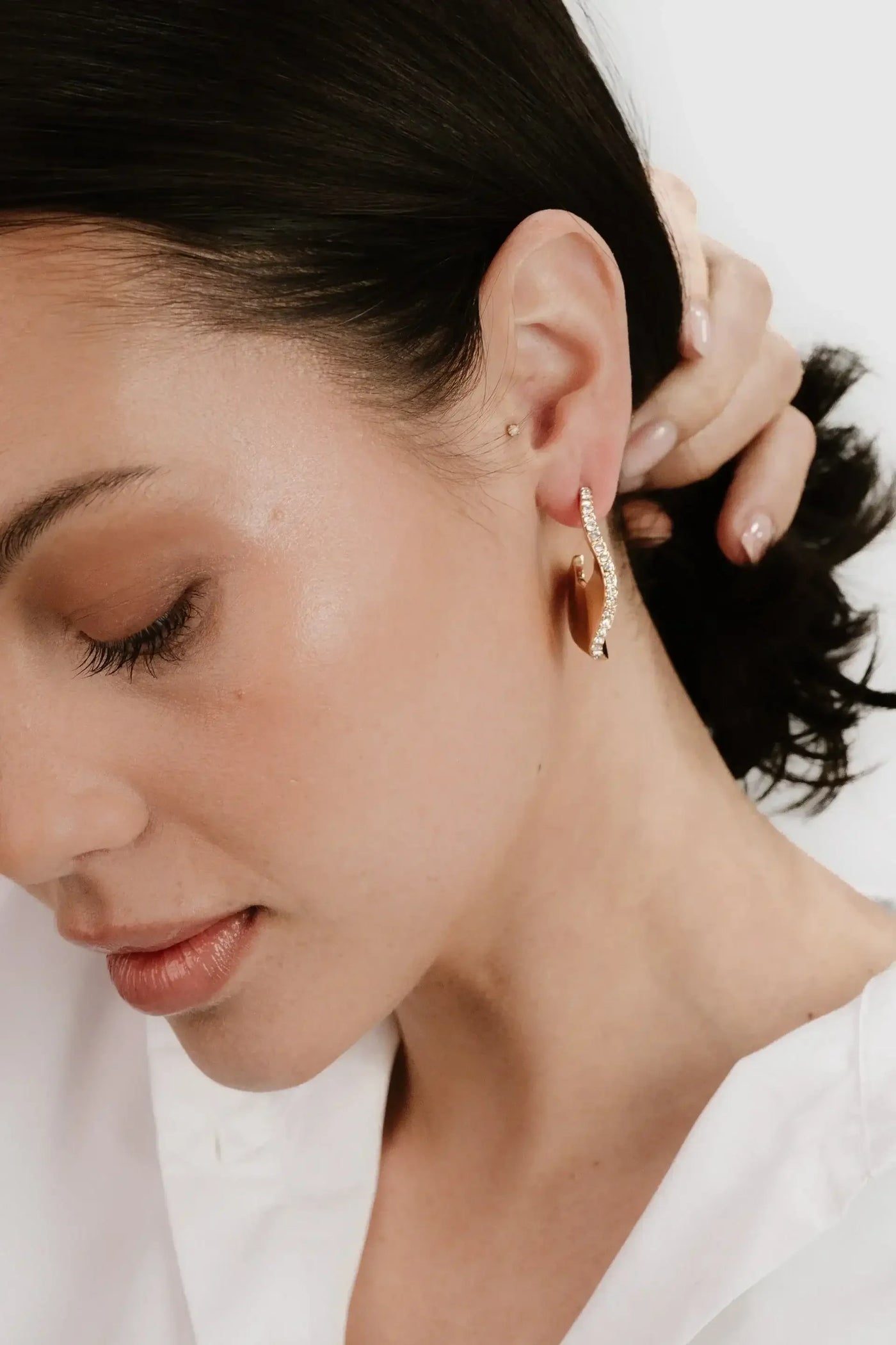 A woman with dark hair touches her ear, where she wears a stylish, curved earring adorned with small stones, set against a neutral background.