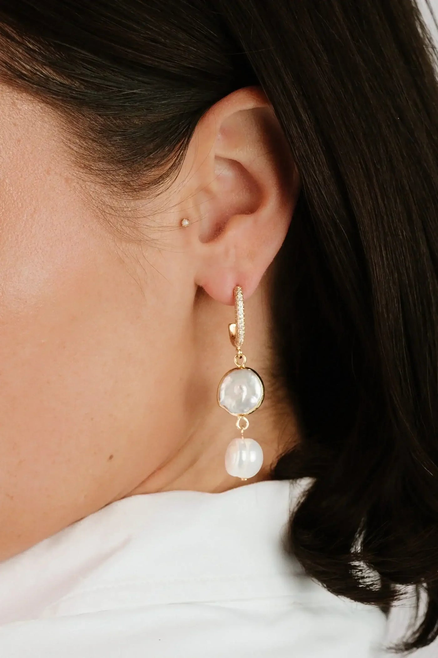 A person wears an elegant gold earring featuring a round pearl and a smaller hanging pearl, set against a smooth, light-colored background and their dark hair, which frames their face.