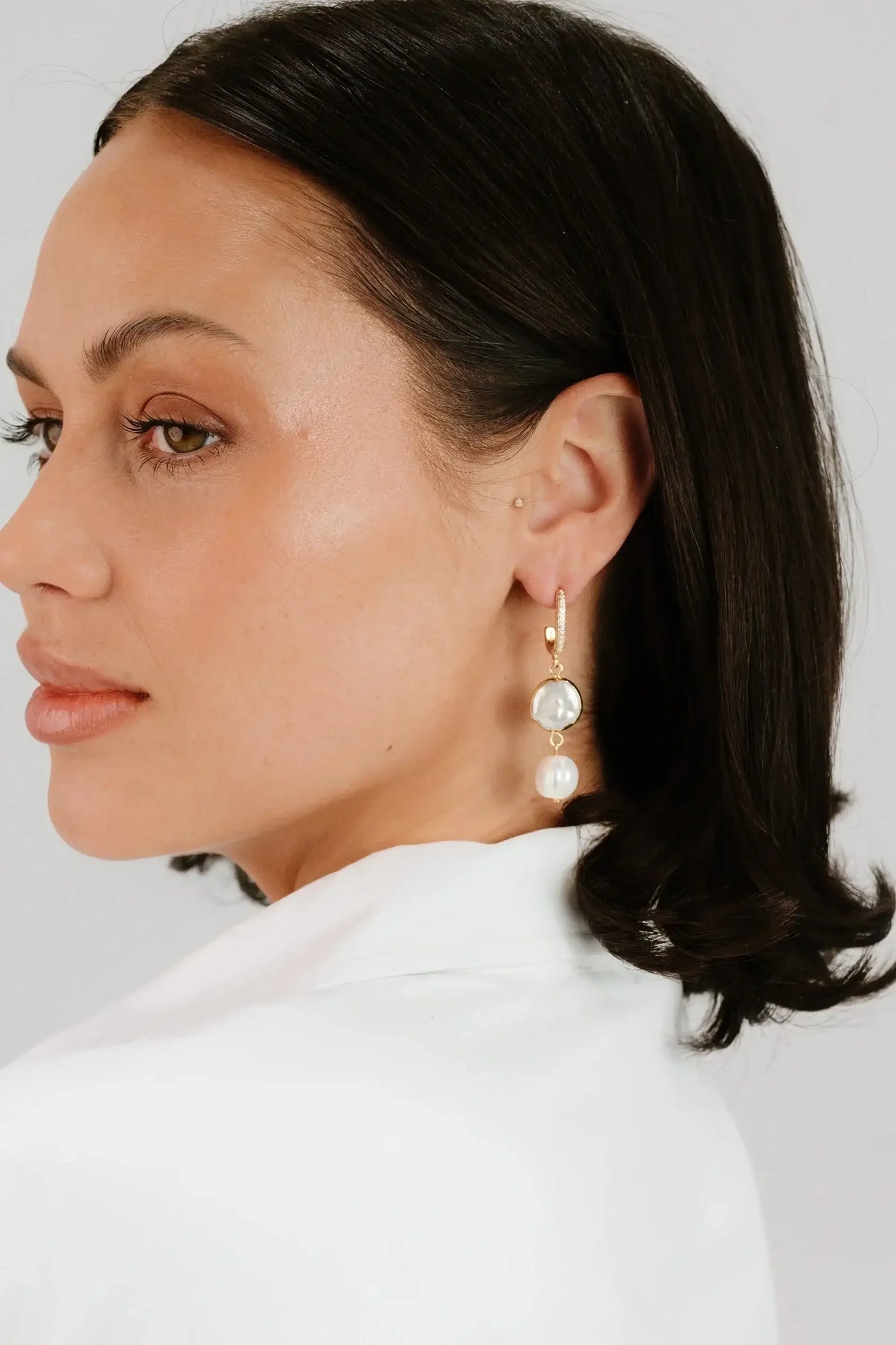 A woman with long, dark hair is wearing elegant earrings featuring a clear stone and a pearl. She is facing slightly to the side against a light background.