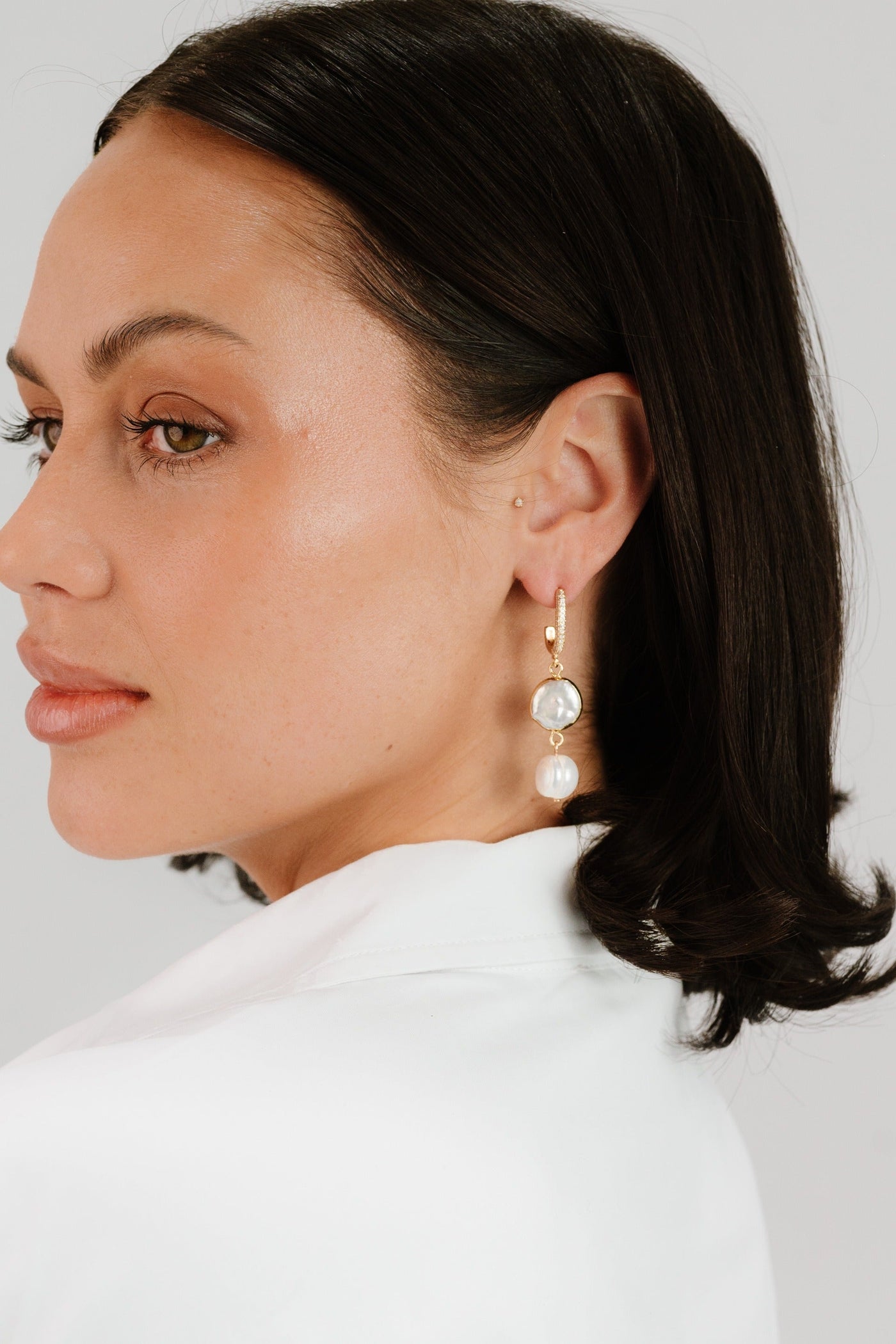 A woman with long, dark hair is wearing elegant earrings featuring a clear stone and a pearl. She is facing slightly to the side against a light background.