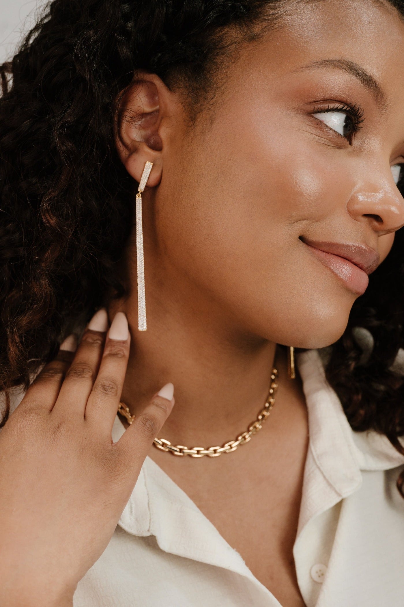 A woman with curly hair is gently touching her neck, showcasing long, sparkling earrings and a gold chain necklace. She wears a light-colored shirt, smiling softly as she turns her head.