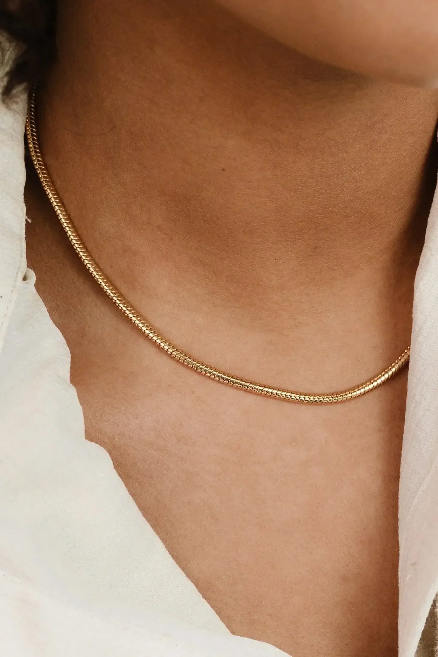 A thin gold necklace rests on a person's neck, gleaming softly. The surrounding skin tone complements the jewelry, which contrasts subtly with a light-colored shirt.
