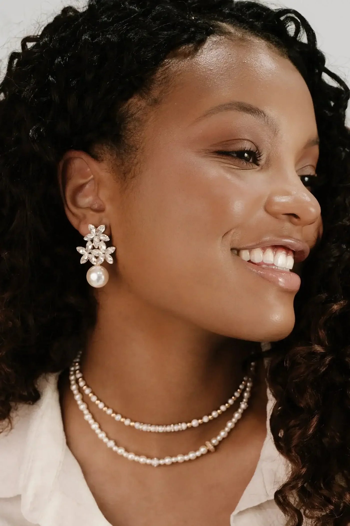 A woman smiles, showcasing large, decorative earrings featuring pearls and gems, paired with layered pearl necklaces, against a soft, neutral background that highlights her accessories.