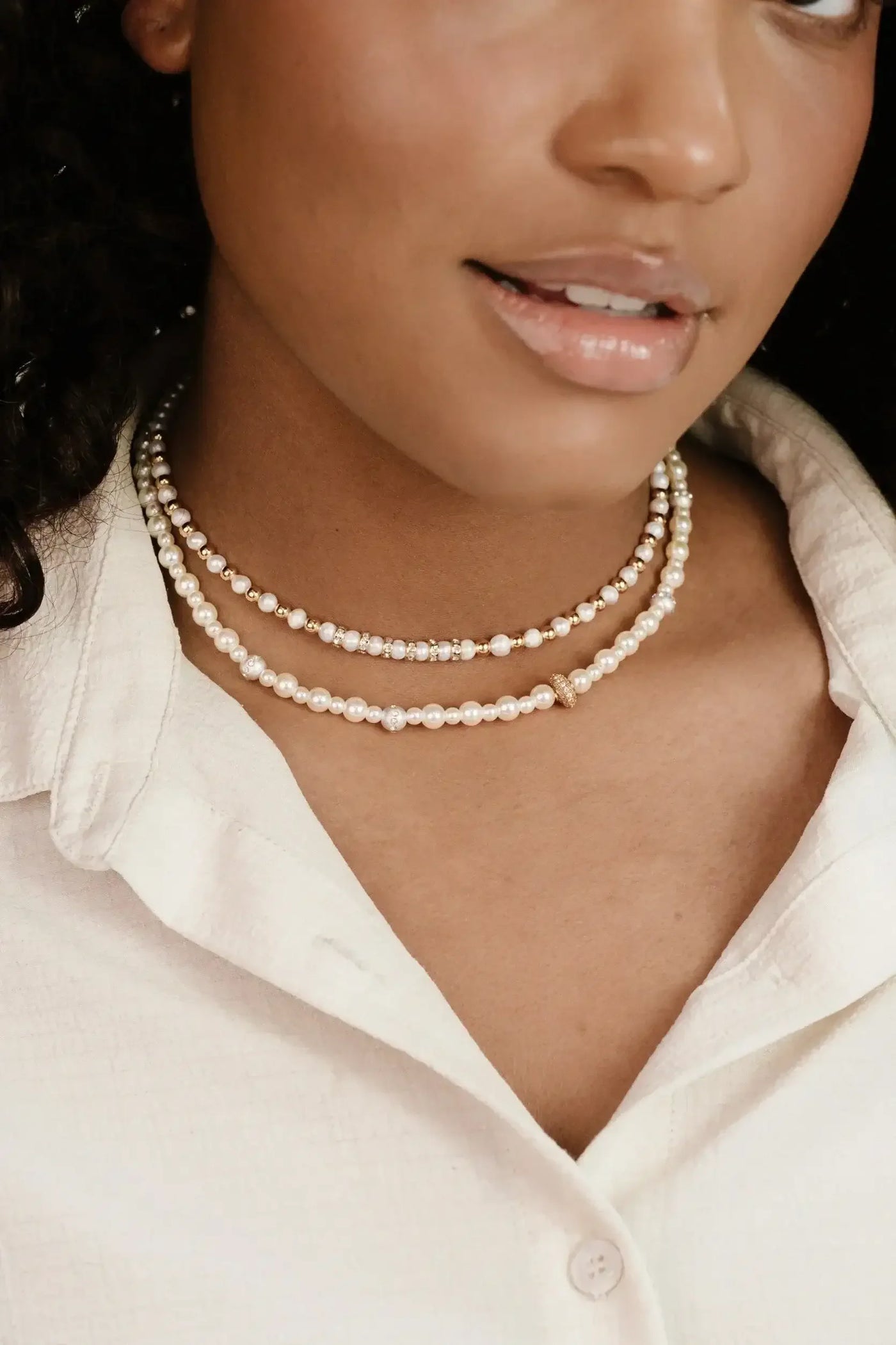 A person wears three layers of pearl necklaces. They smile softly. The individual has curly hair and is dressed in a light-colored, collared shirt, creating a casual, elegant appearance.