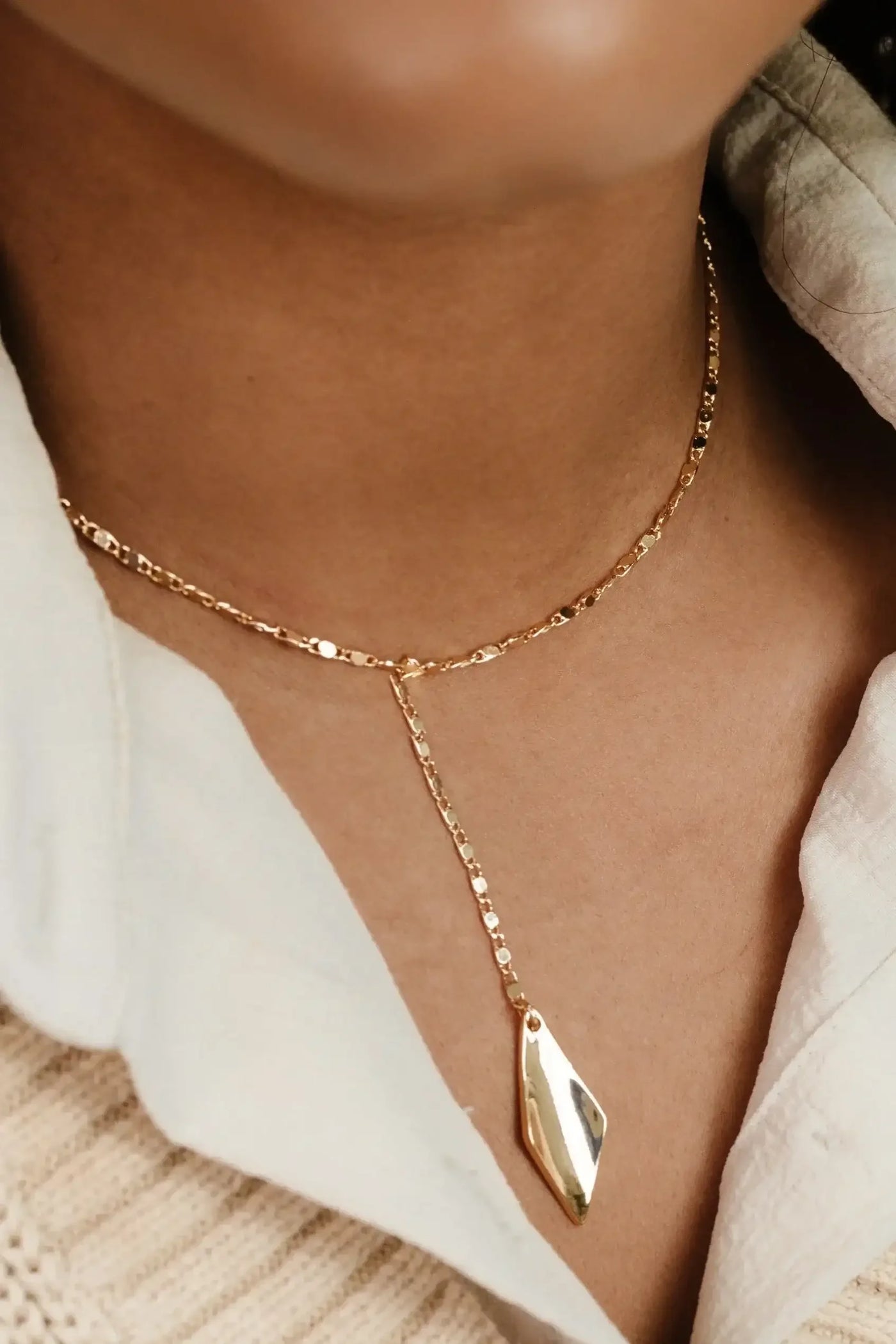 A gold necklace lays on a bare neck, featuring a delicate chain and a geometric pendant hanging down. It's contrasted by the soft fabric of a blouse.
