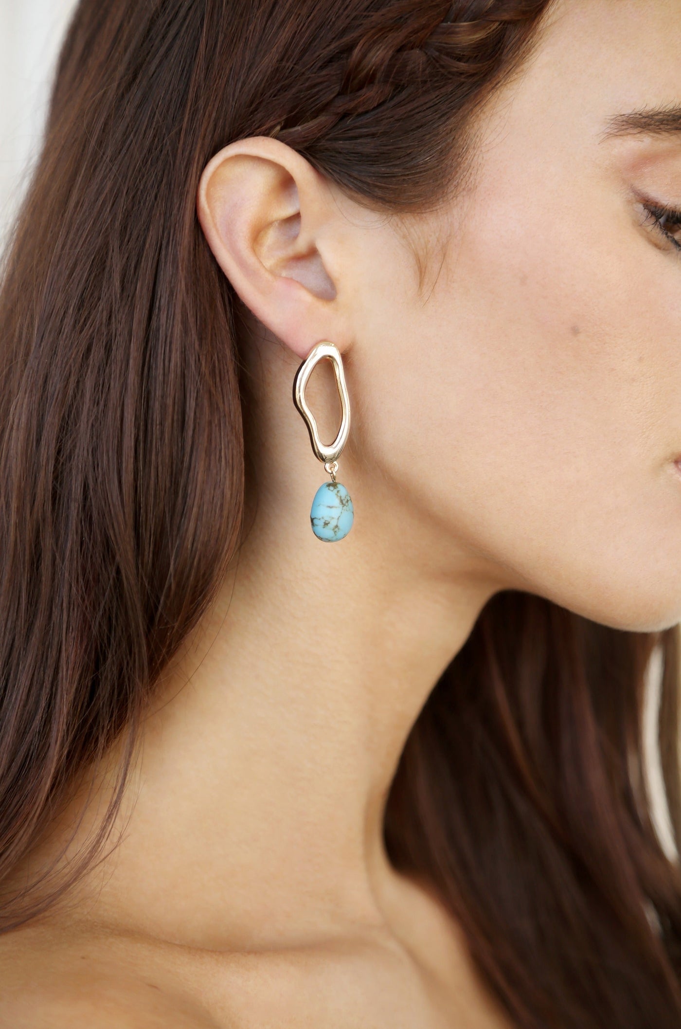 A large, abstract gold earring hangs from a woman's ear, featuring a turquoise stone beneath it. The setting is neutral and soft, highlighting her side profile.