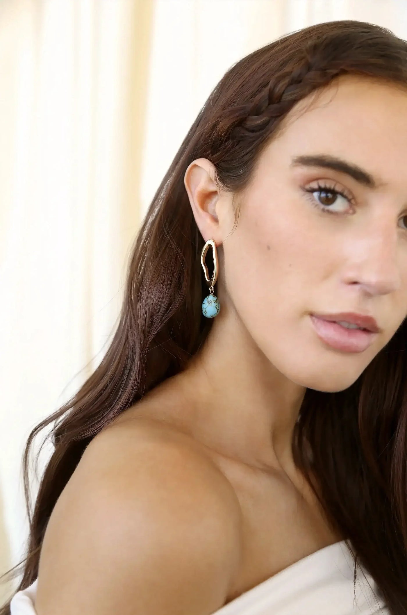 A woman with long, dark hair wears a stylish earring featuring a gold curve and a turquoise drop. She poses with a soft expression against a light-colored, blurred background.
