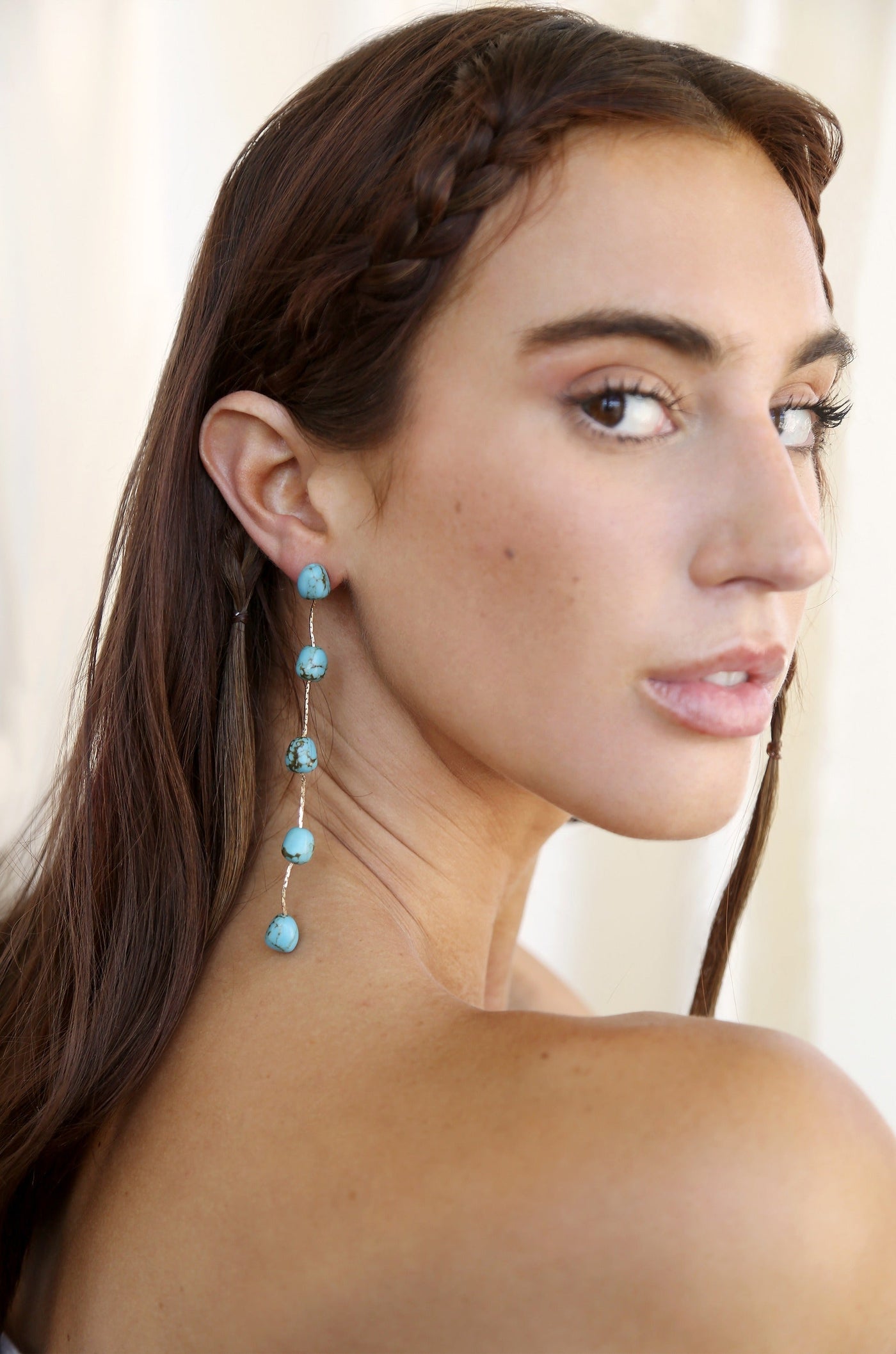 A woman with long, brown hair featuring a braid poses, showcasing a pair of dangling turquoise earrings against a soft, light-colored background. Her expression is serene and thoughtful.