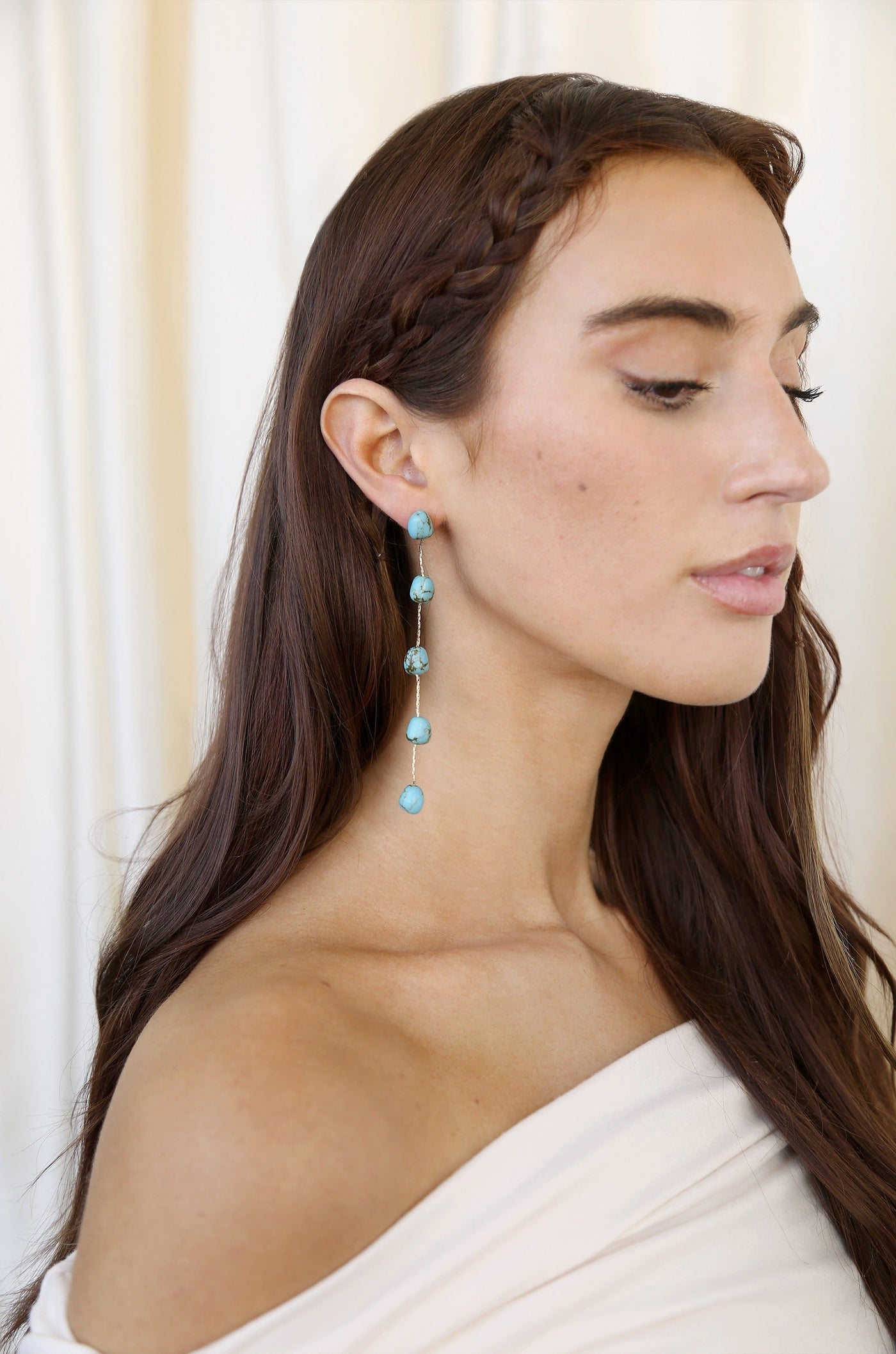 A woman with long, dark hair and a braided section is wearing dangling earrings made of turquoise stones. She is positioned against a soft, light-colored background.