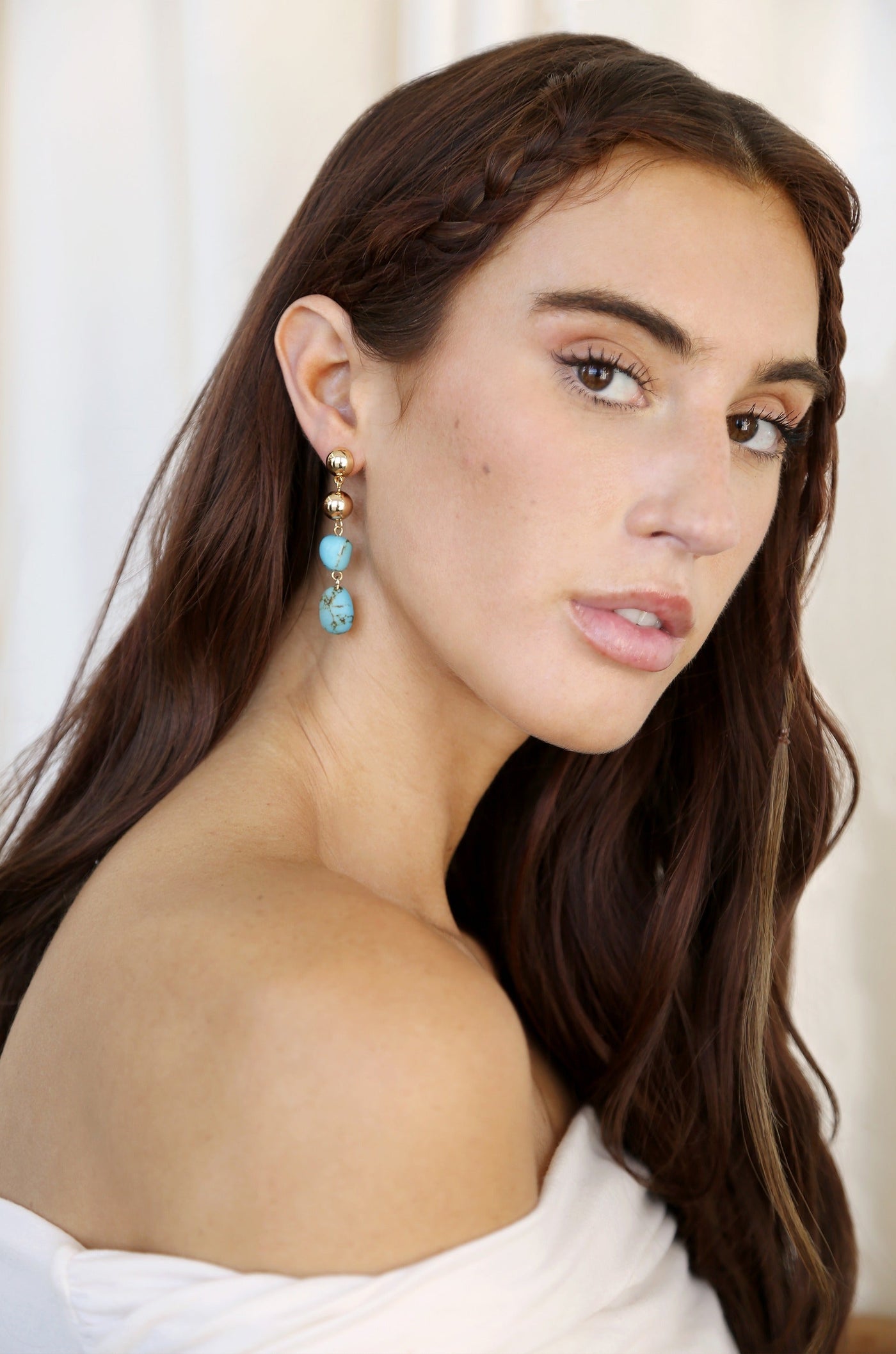 A woman with long, brown hair, adorned with a braid, turns her head slightly, showcasing elegant earrings with turquoise and gold accents against a softly lit, neutral background.