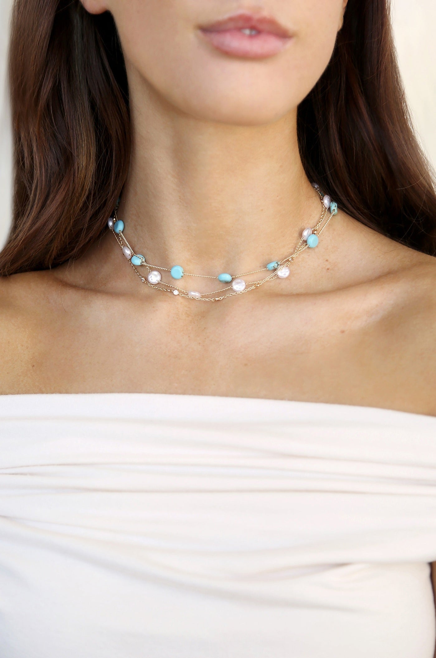 A layered necklace adorned with turquoise and pearl beads rests on a woman's neck, complementing her off-shoulder top in a soft, neutral setting. Her expression is serene, enhancing the elegance of the jewelry.