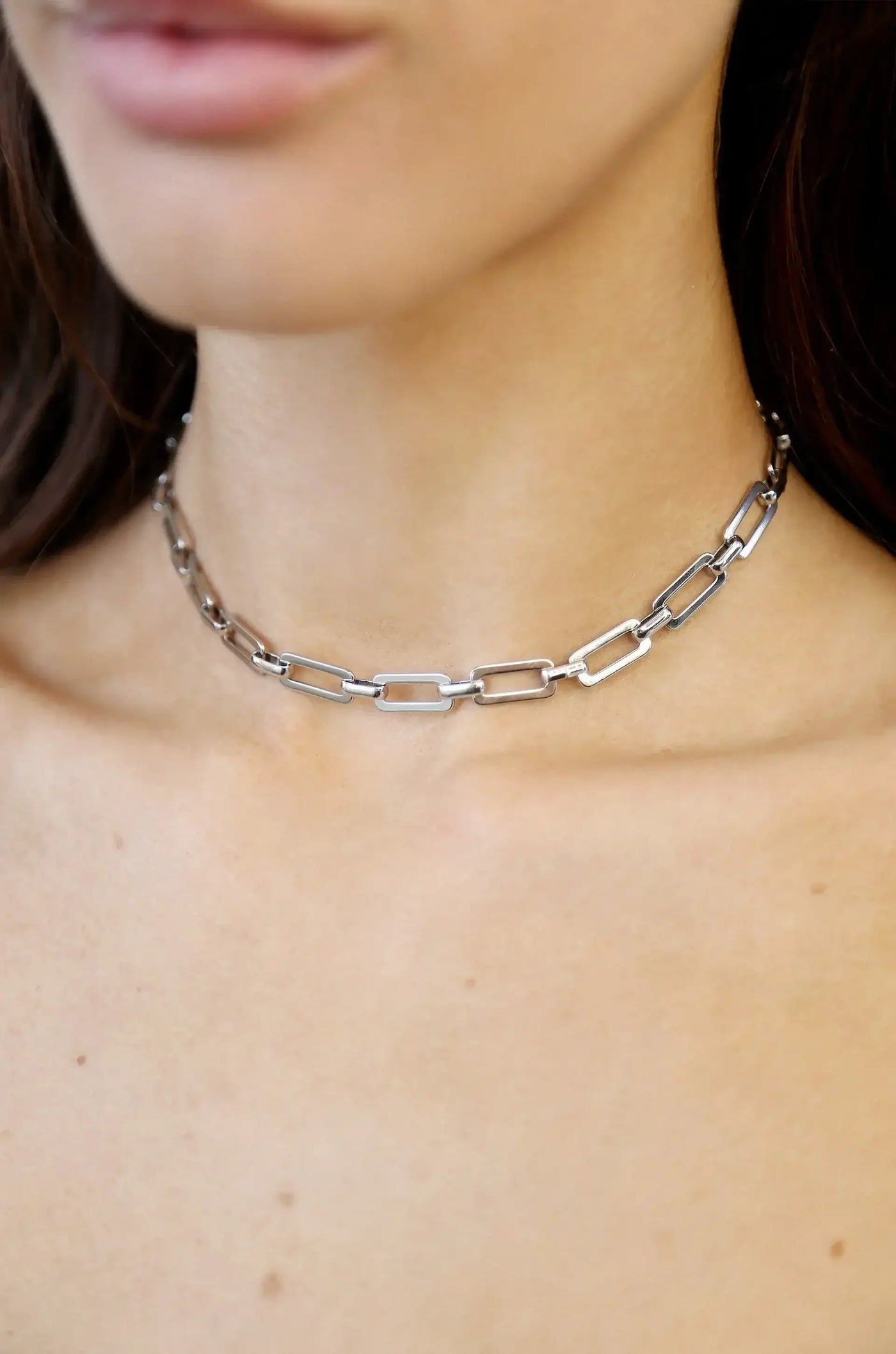 A silver chain necklace with elongated, linked segments rests on the neck of a person, highlighting smooth skin and a subtle collarbone, against a neutral background.