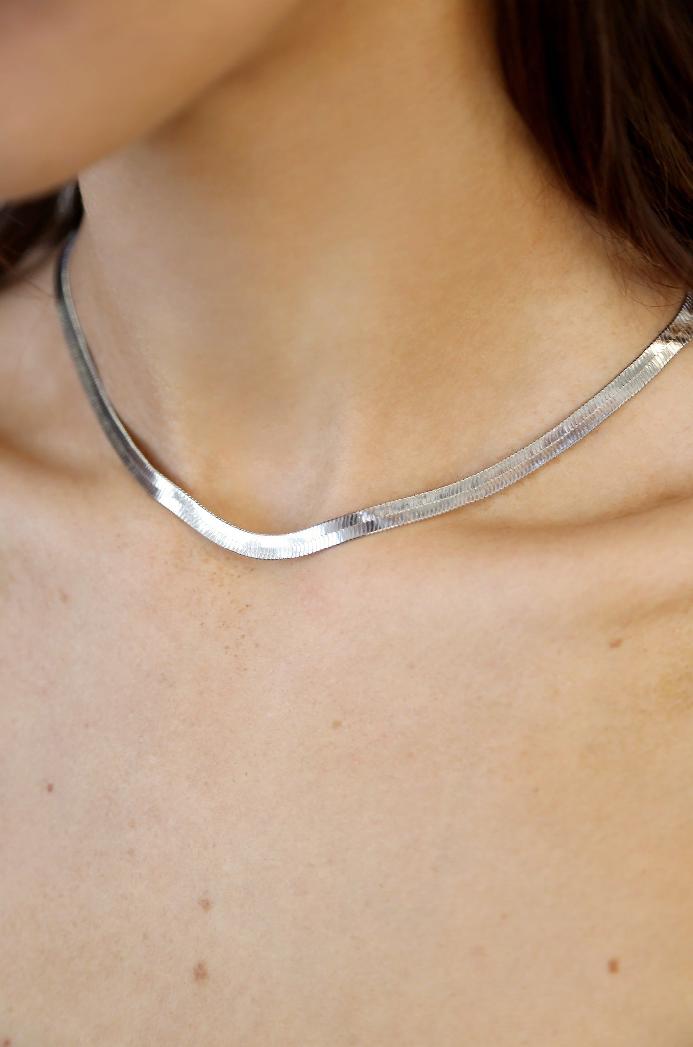 A silver necklace rests elegantly around a person’s neck, reflecting light against their smooth skin, which features subtle freckles in a neutral-toned setting.