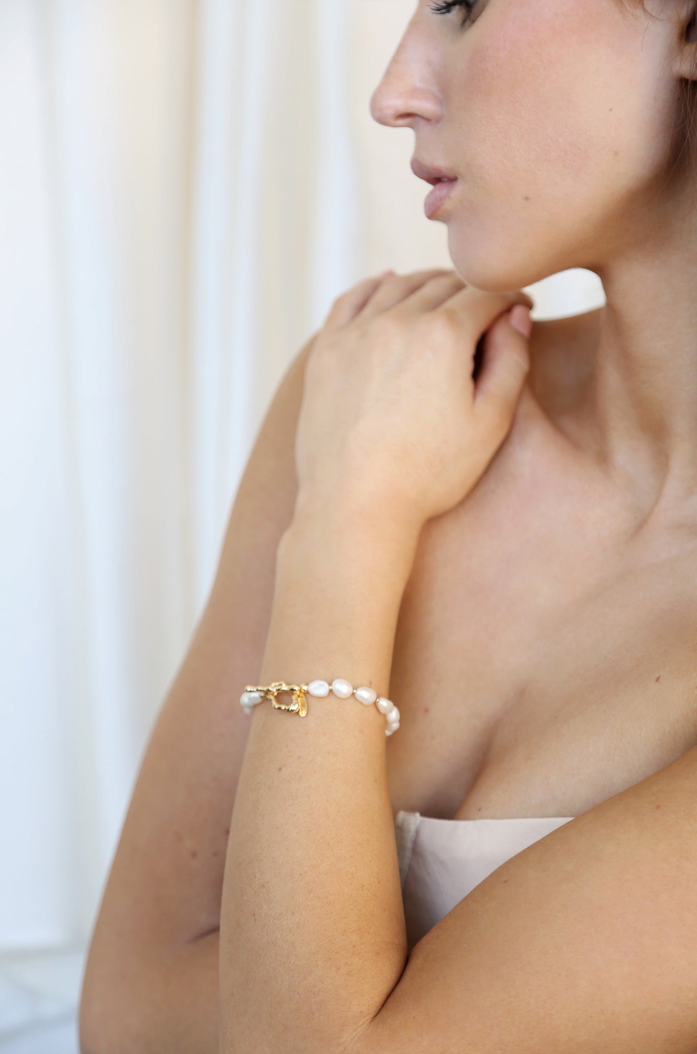 A close-up of a woman's arm adorned with a pearl bracelet featuring a gold clasp. She gently touches her collarbone, set against a soft, light background.