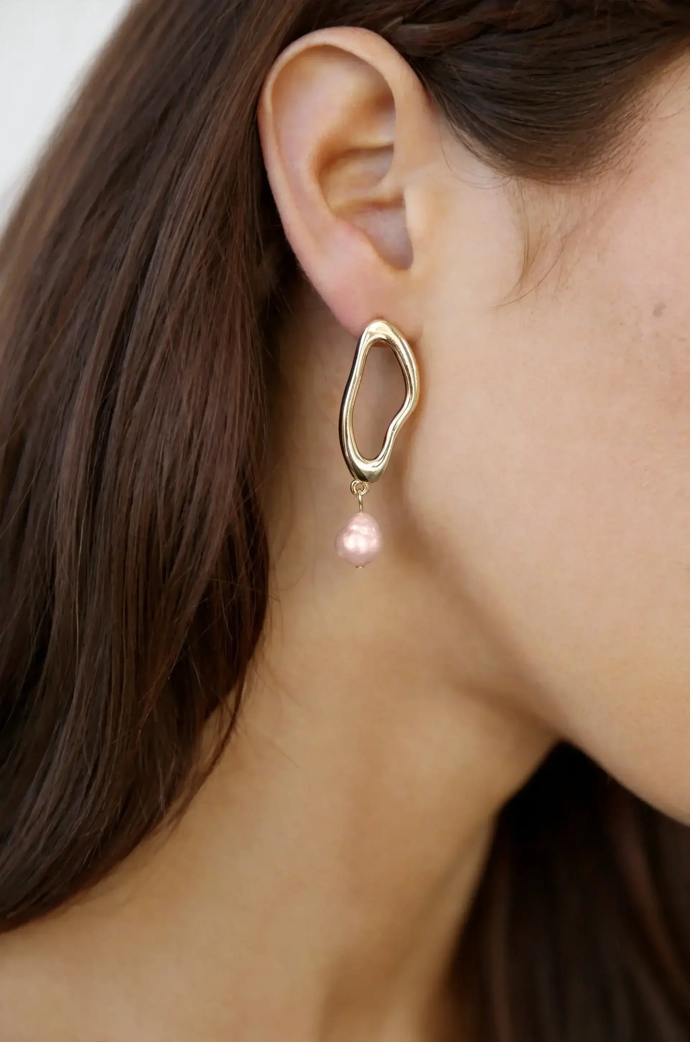 A unique gold earring featuring an abstract shape hangs from a woman's ear. It has a pink pearl dangling below it, set against a softly blurred background of hair and skin.