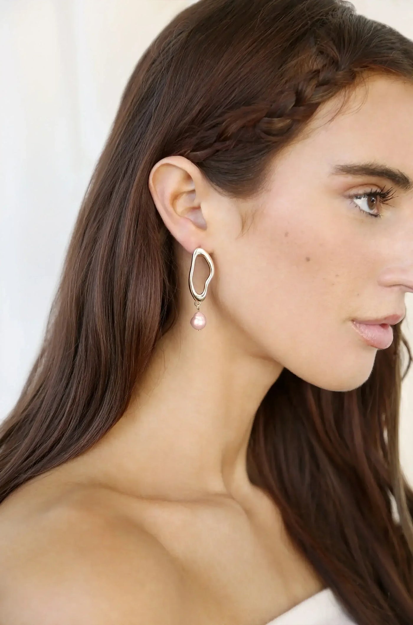 A young woman is wearing a contemporary earring featuring a textured silver outline and a pink pearl drop. Her hair is long and straight, with a braid on one side. The background is softly blurred.