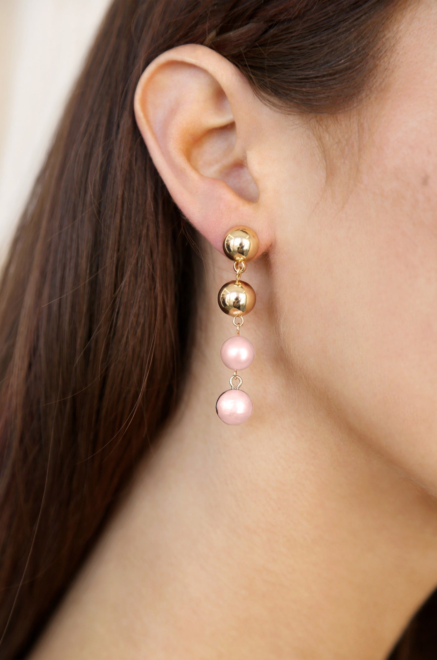 A pair of earrings features a gold stud at the top, dangling two pink pearls below. The earrings are adorned on the ear of a person with long brown hair, set against a neutral background.