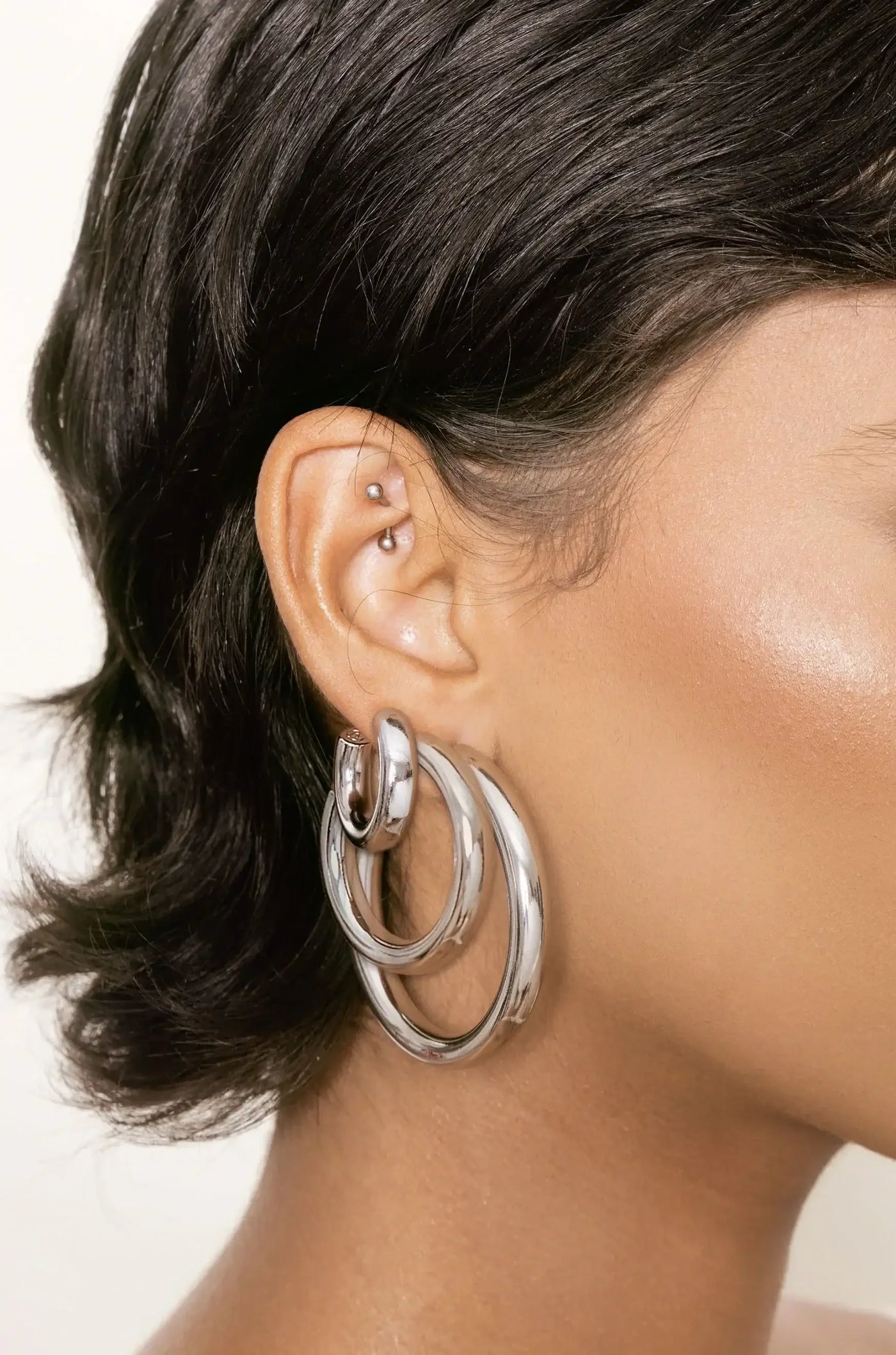 A large, shiny silver hoop earring hangs from the individual's ear. The person has short, dark hair and smooth, tanned skin, with an ear piercing visible above the earring.