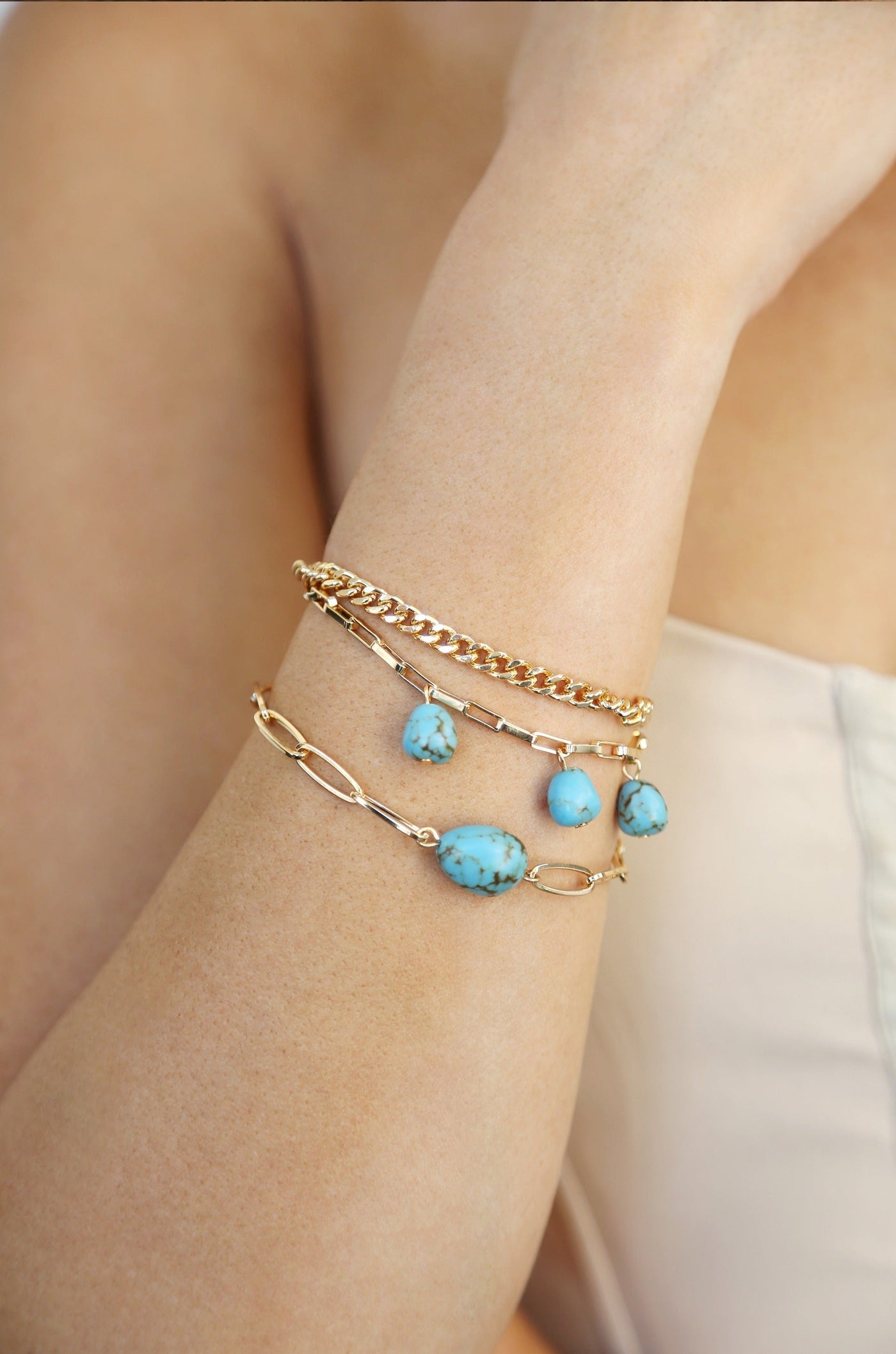 A gold bracelet with turquoise stones adorns a woman's wrist. The skin is smooth and lightly tanned, while the background is a soft neutral color, enhancing the jewelry's elegance.