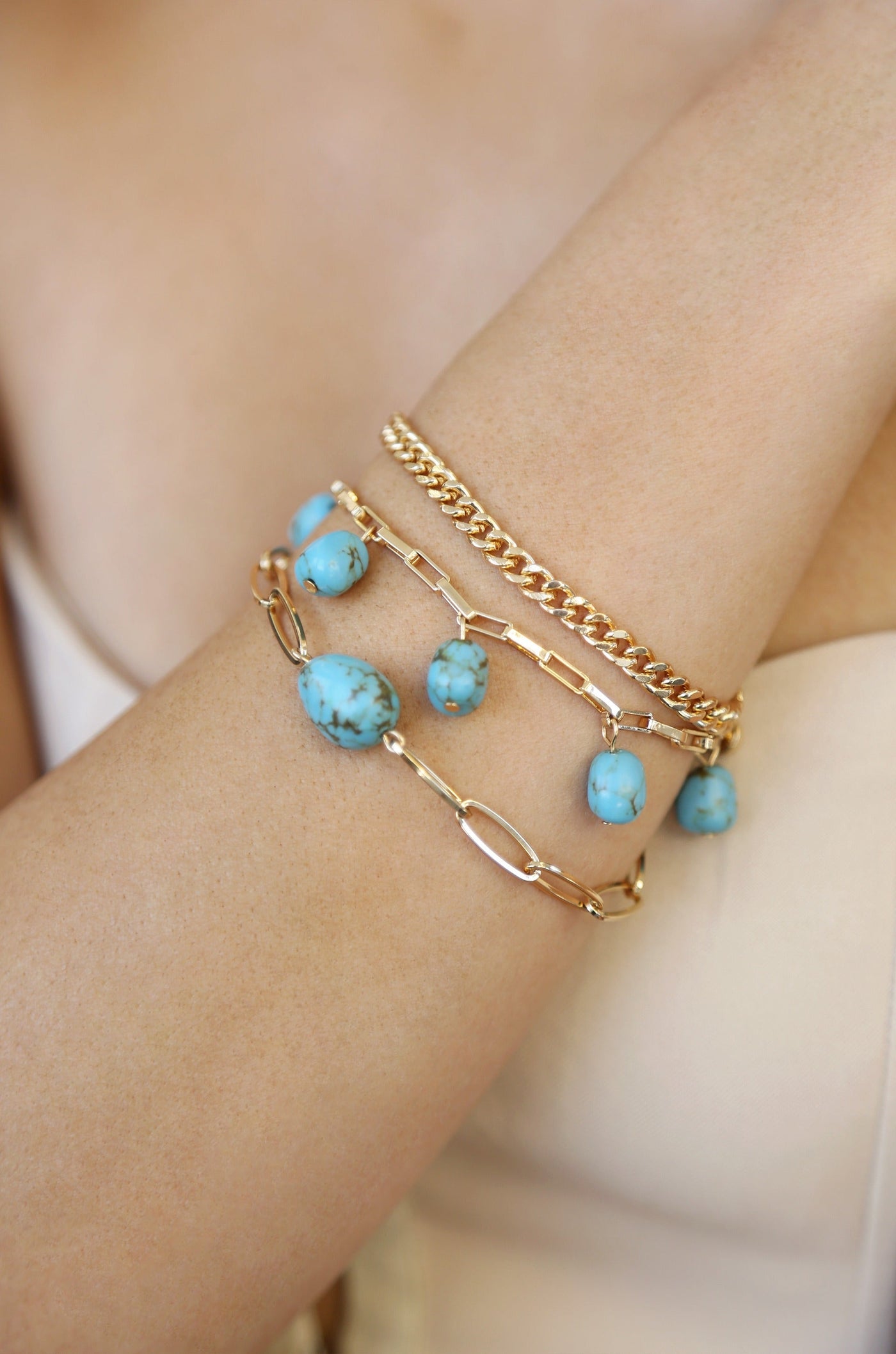 A close-up of a wrist displaying three bracelets: one with turquoise stones and gold links, one gold chain, and one gold chain with varying links, set against a light beige background.