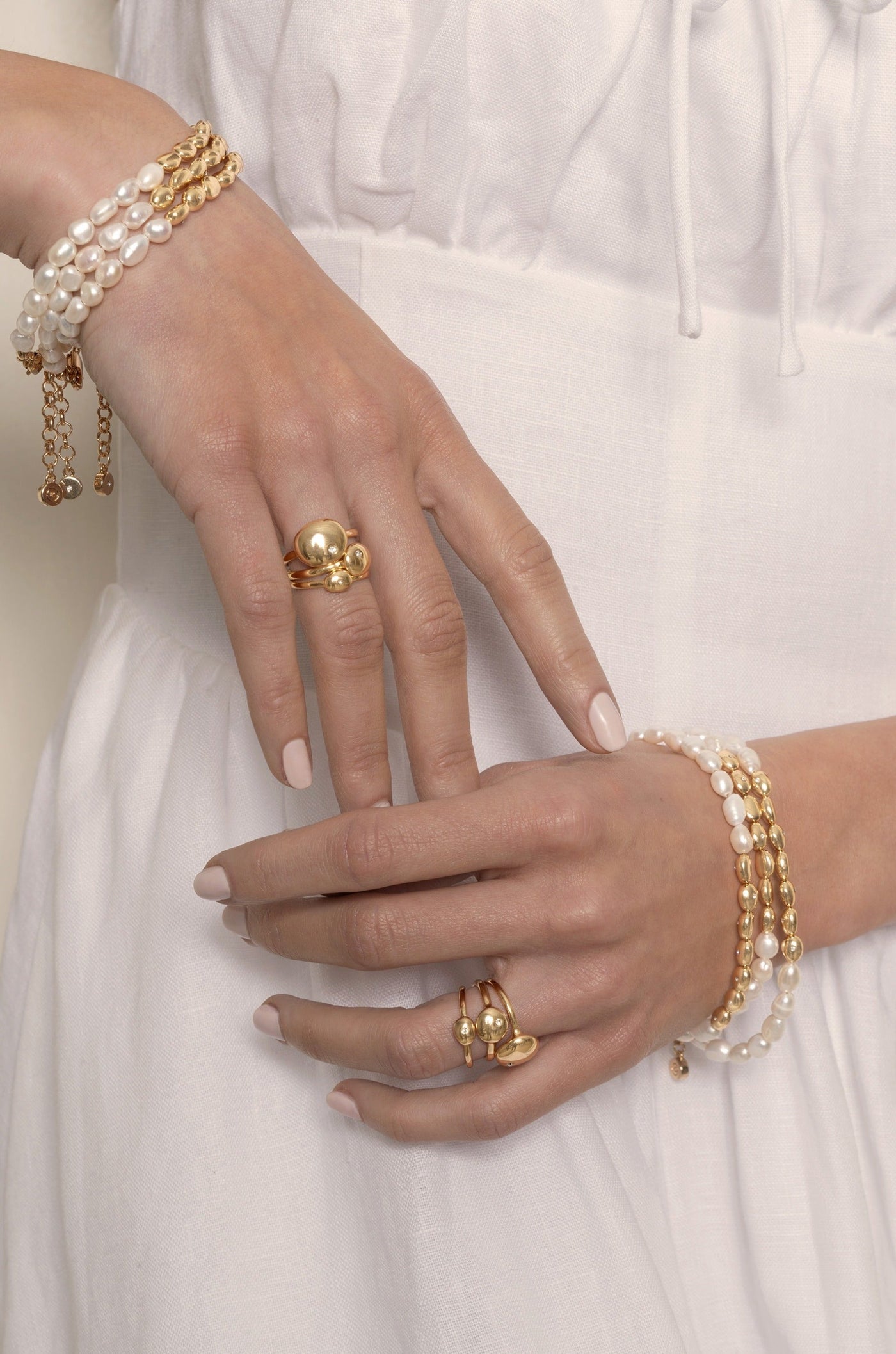 A hand adorned with multiple gold and pearl bracelets gestures softly, showcasing a gold ring set on its fingers, all against a soft, white fabric backdrop.