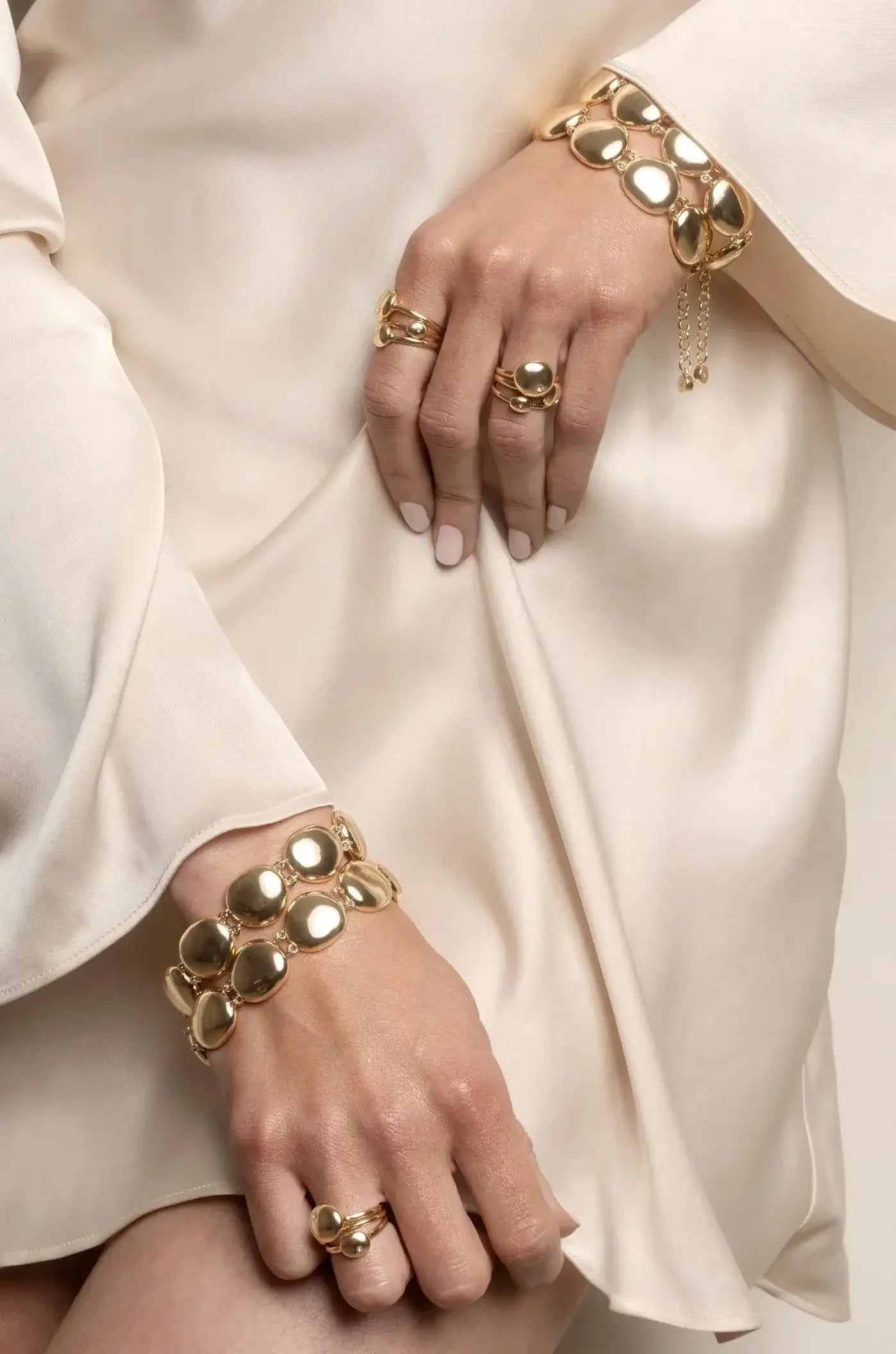 Gold jewelry, including rings and a bracelet, adorns elegantly manicured hands resting on a soft, pale fabric. The overall context suggests a refined fashion presentation.