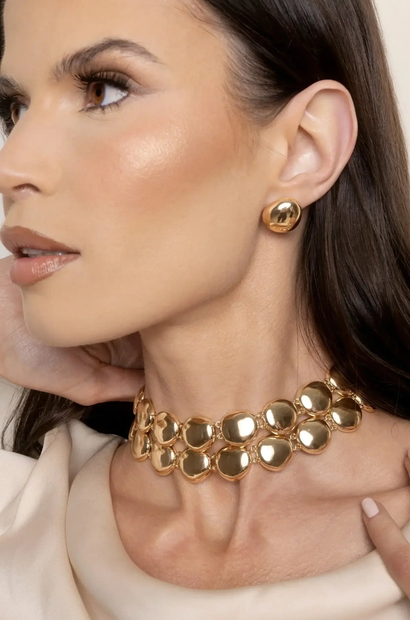 A woman displays golden jewelry, including a bold choker and matching earrings. She gently touches her neck, exuding elegance against a soft, neutral background. Her makeup enhances her features.