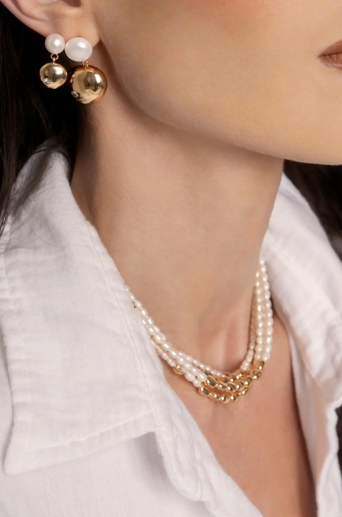 A close-up of a woman's neck showcases elegant jewelry: pearl and gold earrings complement a layered pearl necklace, all against a white collared shirt, emphasizing a chic, sophisticated style.