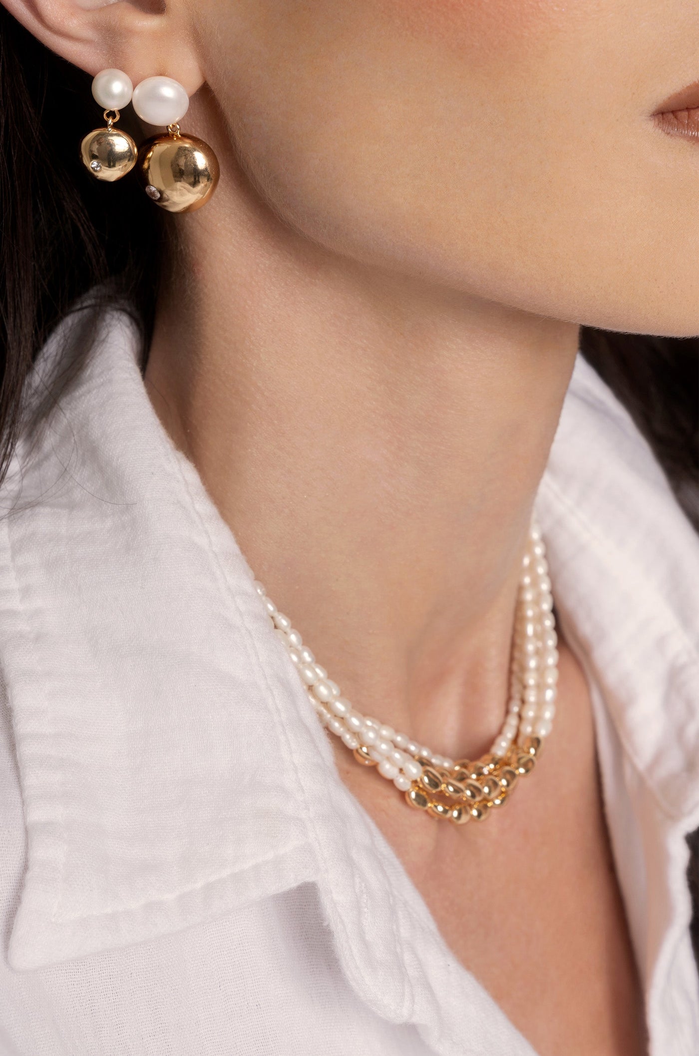 A close-up of a woman's neck showcases elegant jewelry: pearl and gold earrings complement a layered pearl necklace, all against a white collared shirt, emphasizing a chic, sophisticated style.