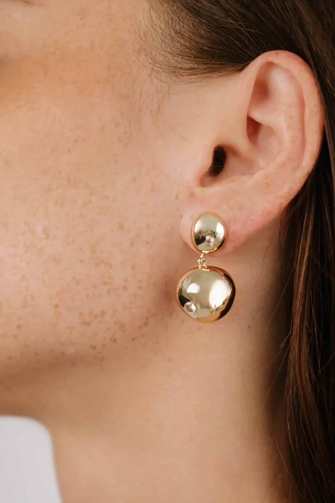 A person's ear showcases a pair of gold earrings—one round and one elongated—reflecting light. The focus is on the jewelry against their lightly freckled skin and brown hair.