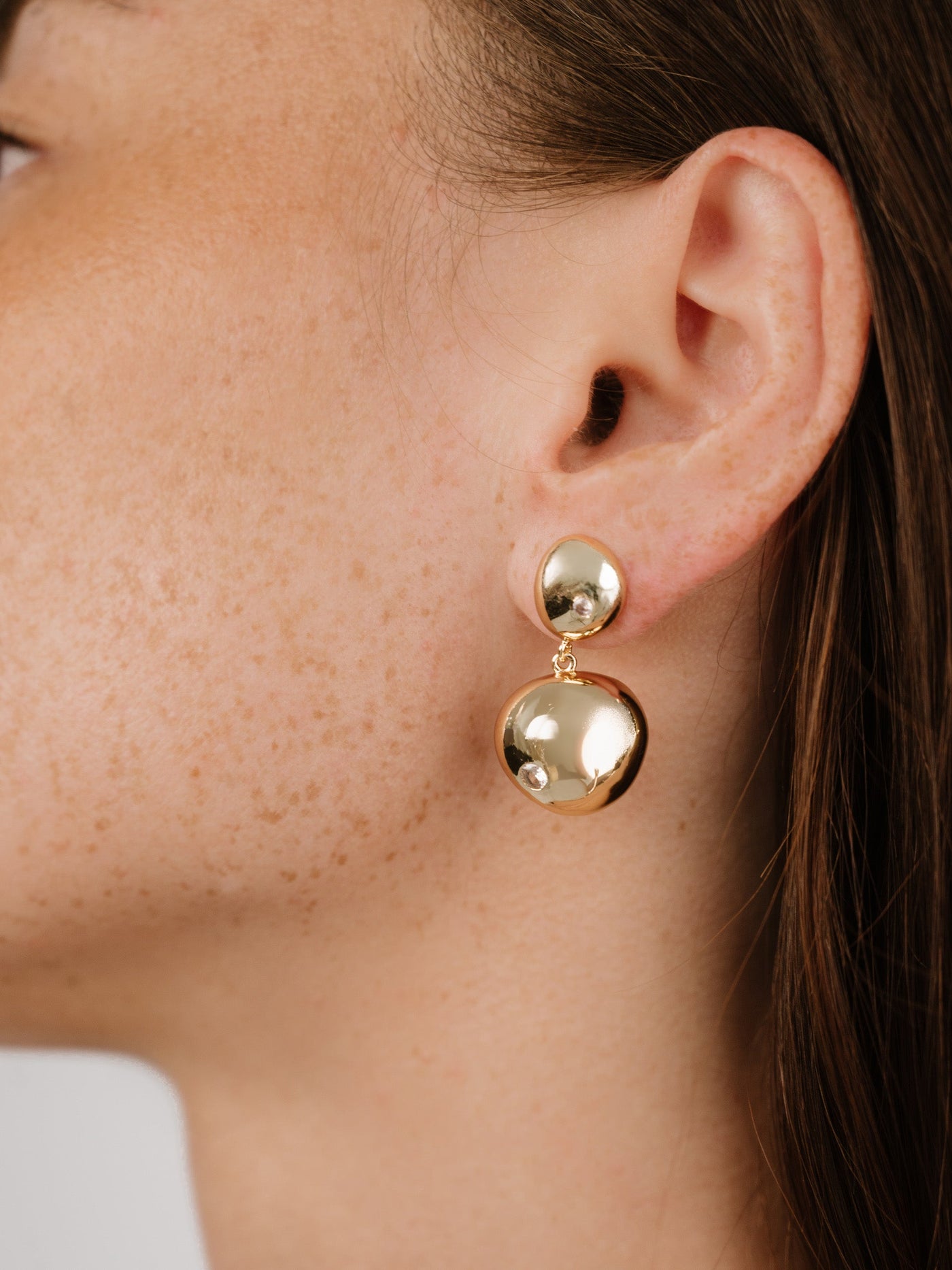 A person's ear showcases a pair of gold earrings—one round and one elongated—reflecting light. The focus is on the jewelry against their lightly freckled skin and brown hair.