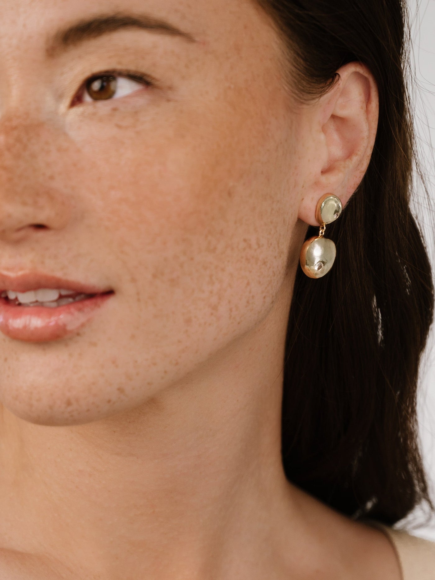 A close-up of a woman’s face showcases her wearing a pair of elegant earrings. She smiles softly, surrounded by a neutral background that highlights her features and accessories.