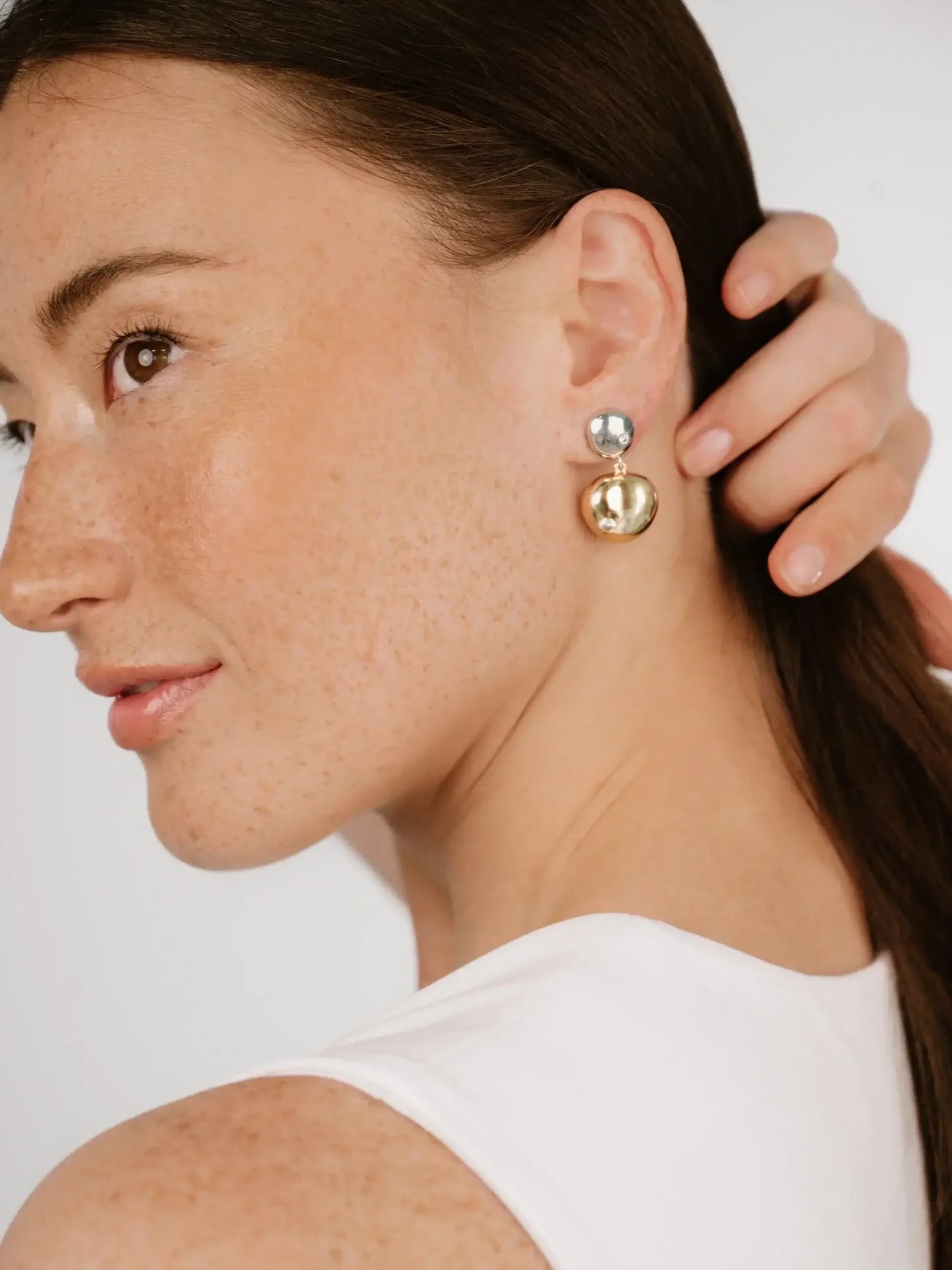 A woman with freckles gently touches her hair while showcasing elegant earrings with a silver top and a gold bottom. The background is neutral and soft, enhancing her features.