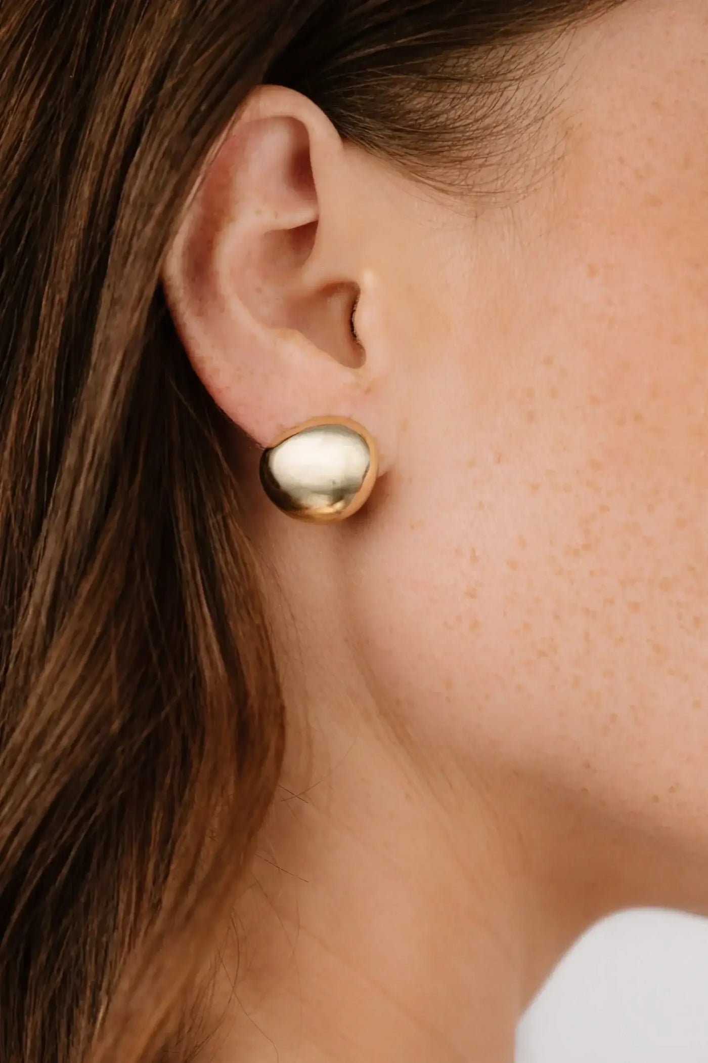 A gold-colored earrings is worn on the ear of a person with light brown hair, showcasing the earring’s polished surface against a background of soft, neutral tones.