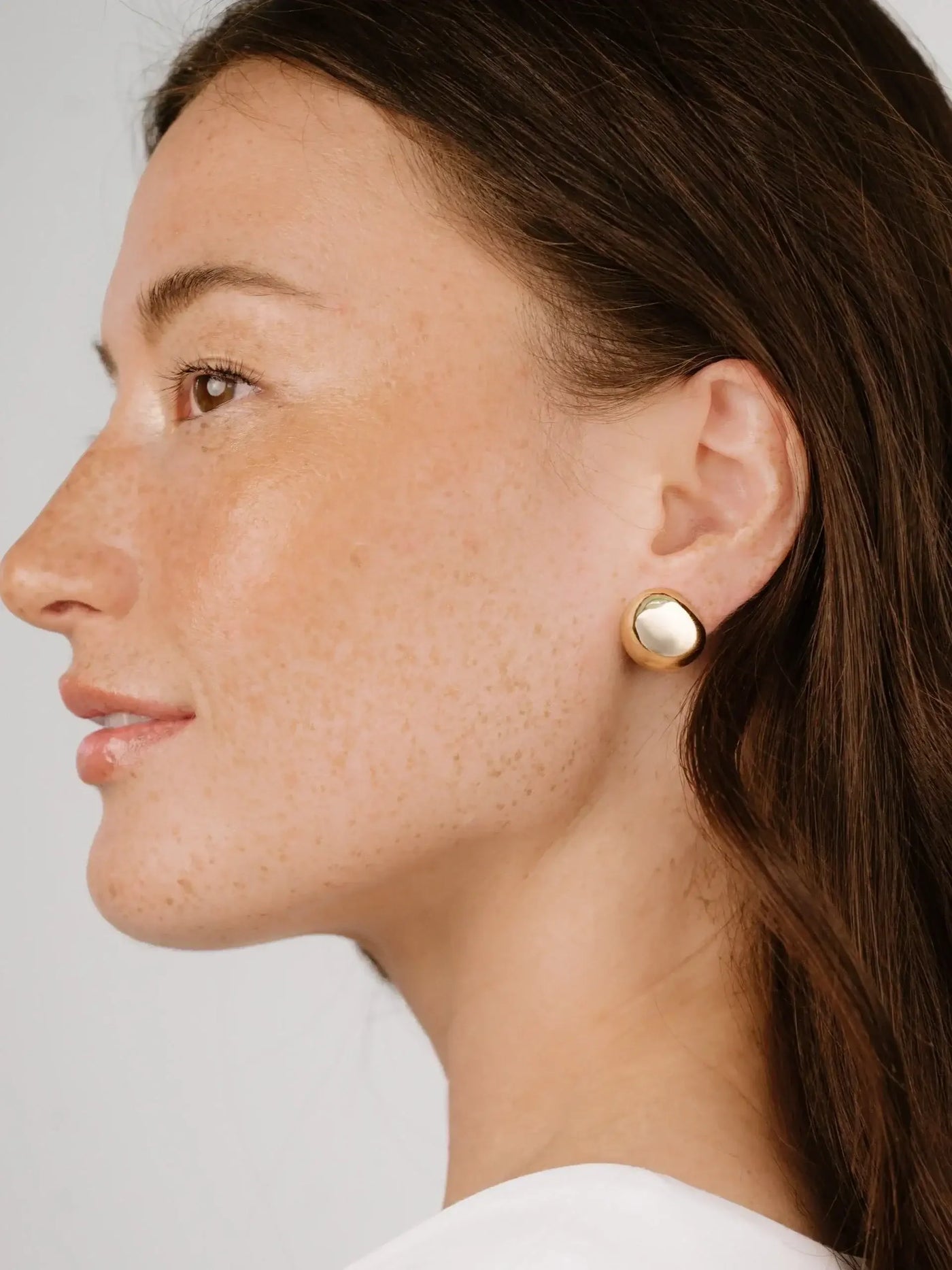 A woman with freckles smiles gently, showcasing a large gold earring in her left ear. Her hair cascades softly around her shoulder against a neutral background.
