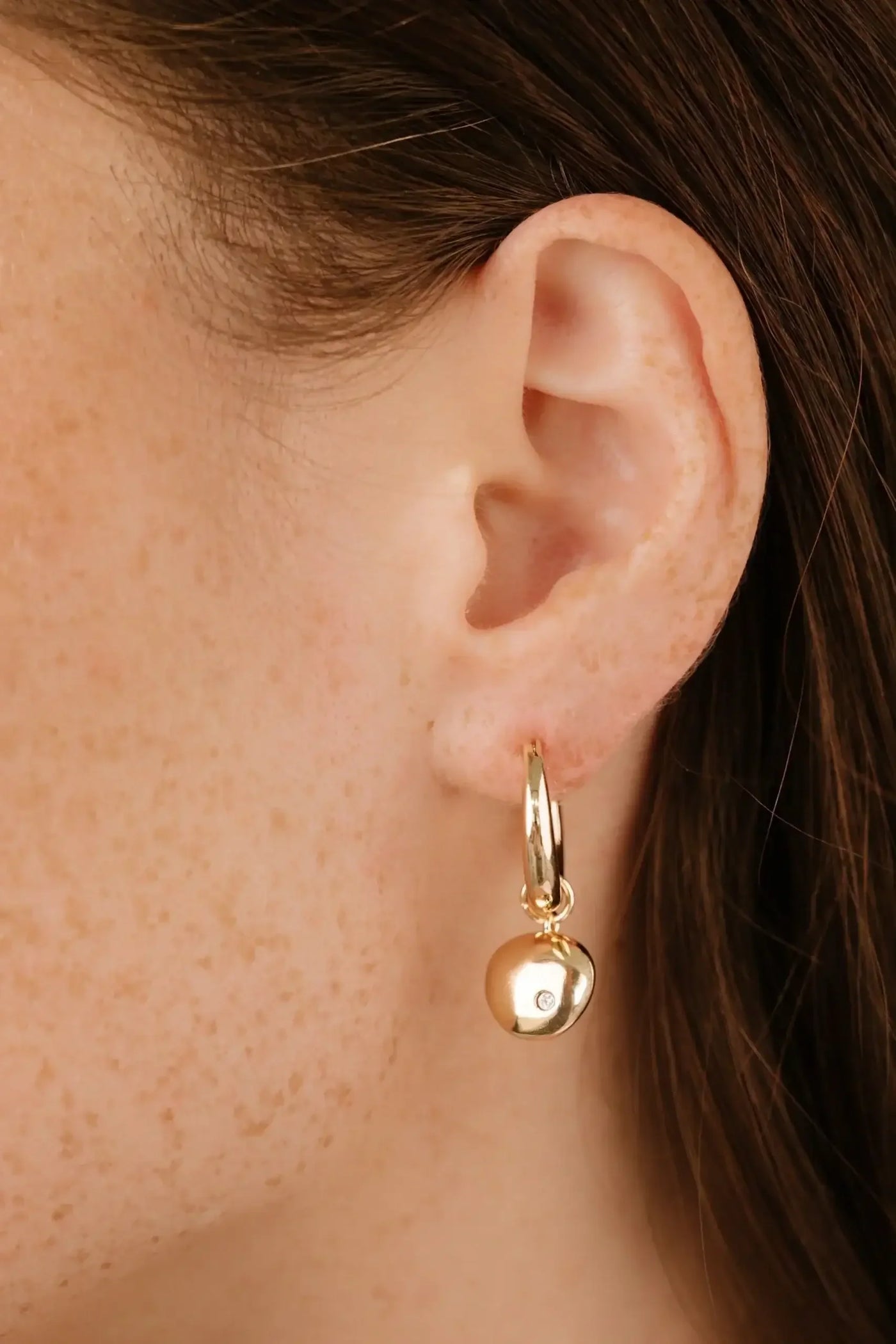 A gold hoop earring with a dangling, round charm is prominently displayed near a woman's ear, showcasing her freckled skin and dark hair in a soft lighting context.