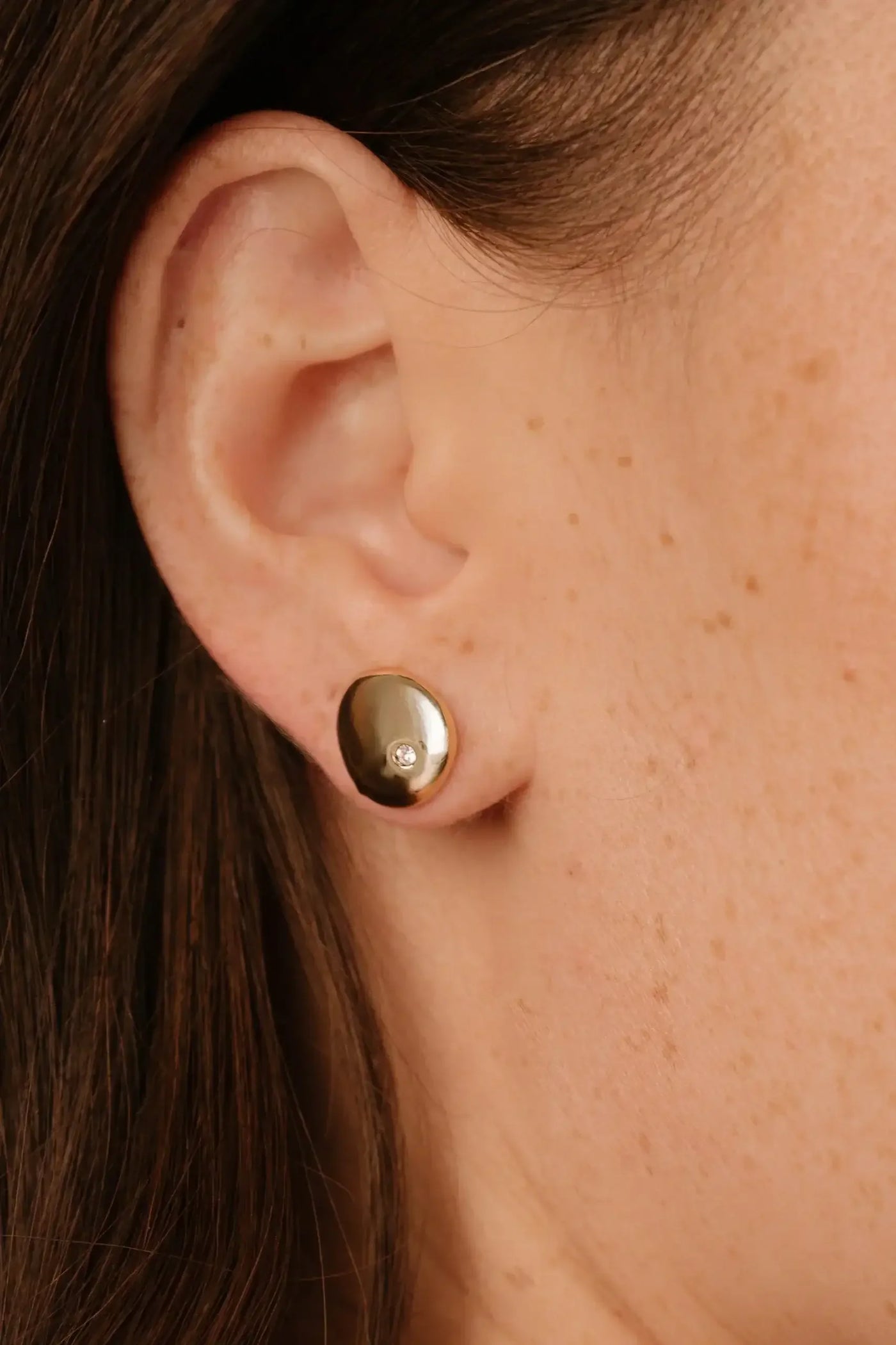 A gold earring is prominently displayed on the ear of a person, featuring a small gemstone. The setting is close-up, highlighting the texture of the skin and earring.