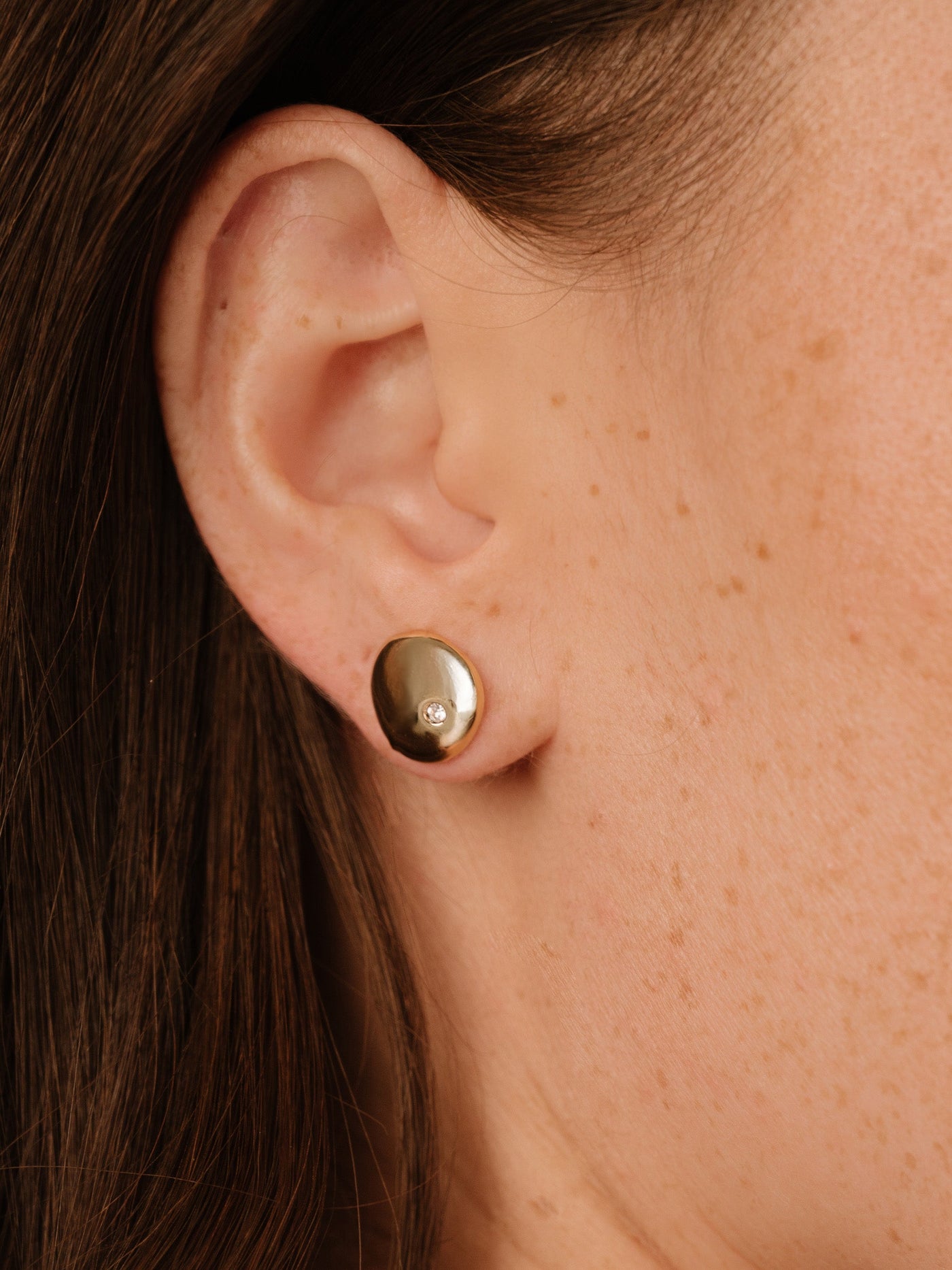 A gold earring is prominently displayed on the ear of a person, featuring a small gemstone. The setting is close-up, highlighting the texture of the skin and earring.