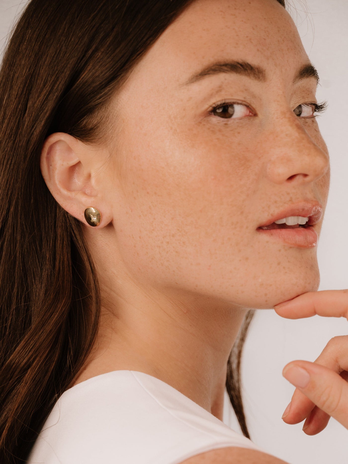 A woman with long brown hair and freckles gently touches her chin while looking slightly over her shoulder, wearing a simple white top and stud earrings against a light background.
