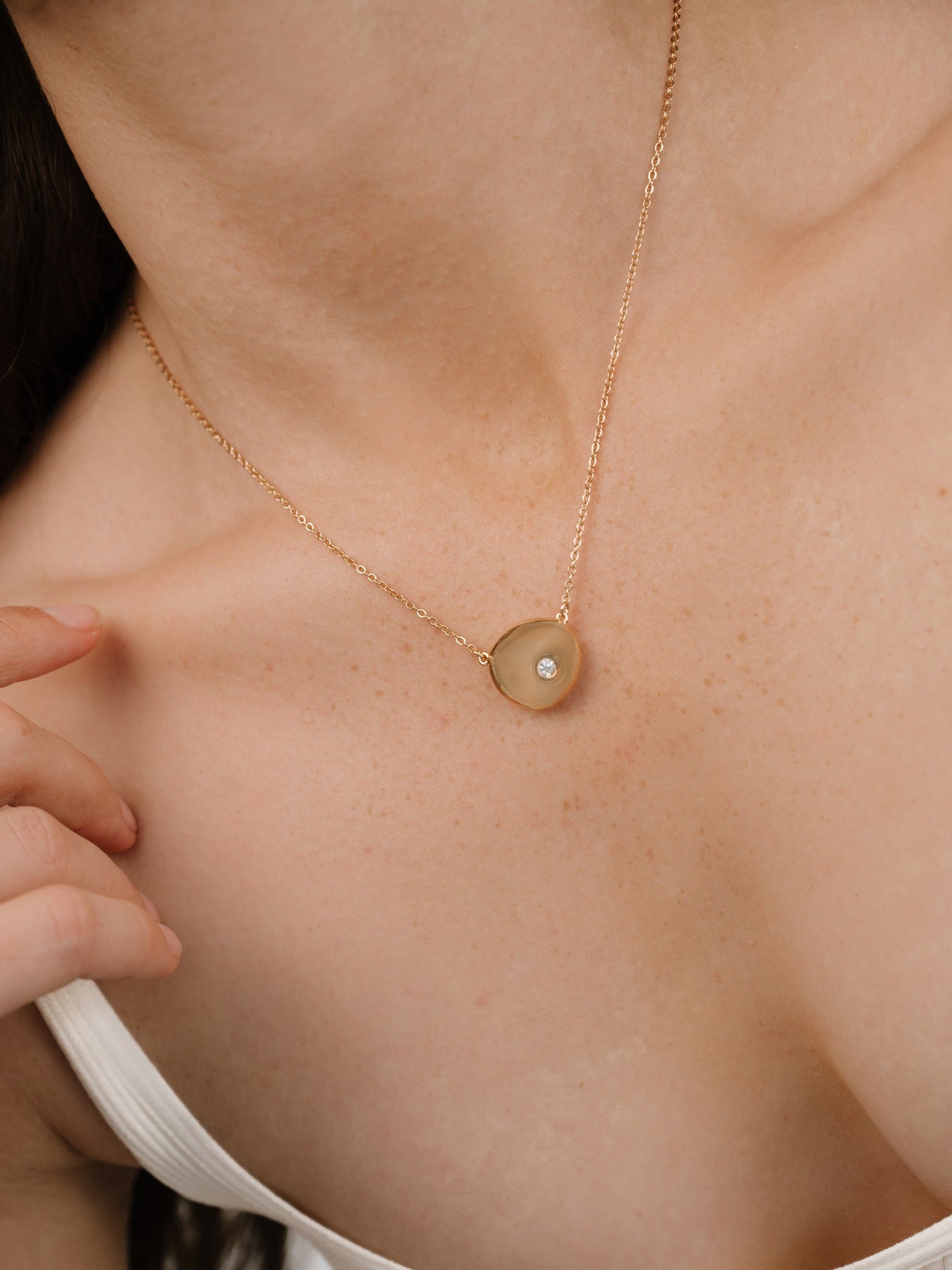 A golden necklace with a circular pendant and a small gemstone rests on a person's collarbone. The individual gently touches the pendant, highlighting its elegance against skin tones.