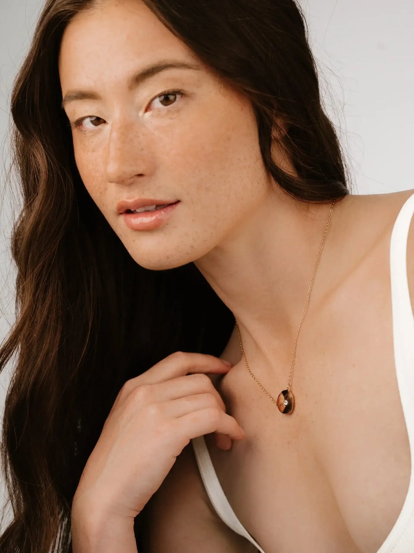 A woman with long, wavy hair poses, her hand gently touching her neckline. She wears a simple necklace, and the background is a soft, neutral color. Natural lighting highlights her features.