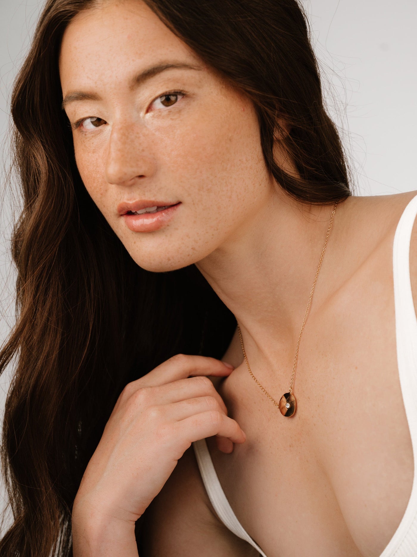 A woman with long, wavy hair poses, her hand gently touching her neckline. She wears a simple necklace, and the background is a soft, neutral color. Natural lighting highlights her features.